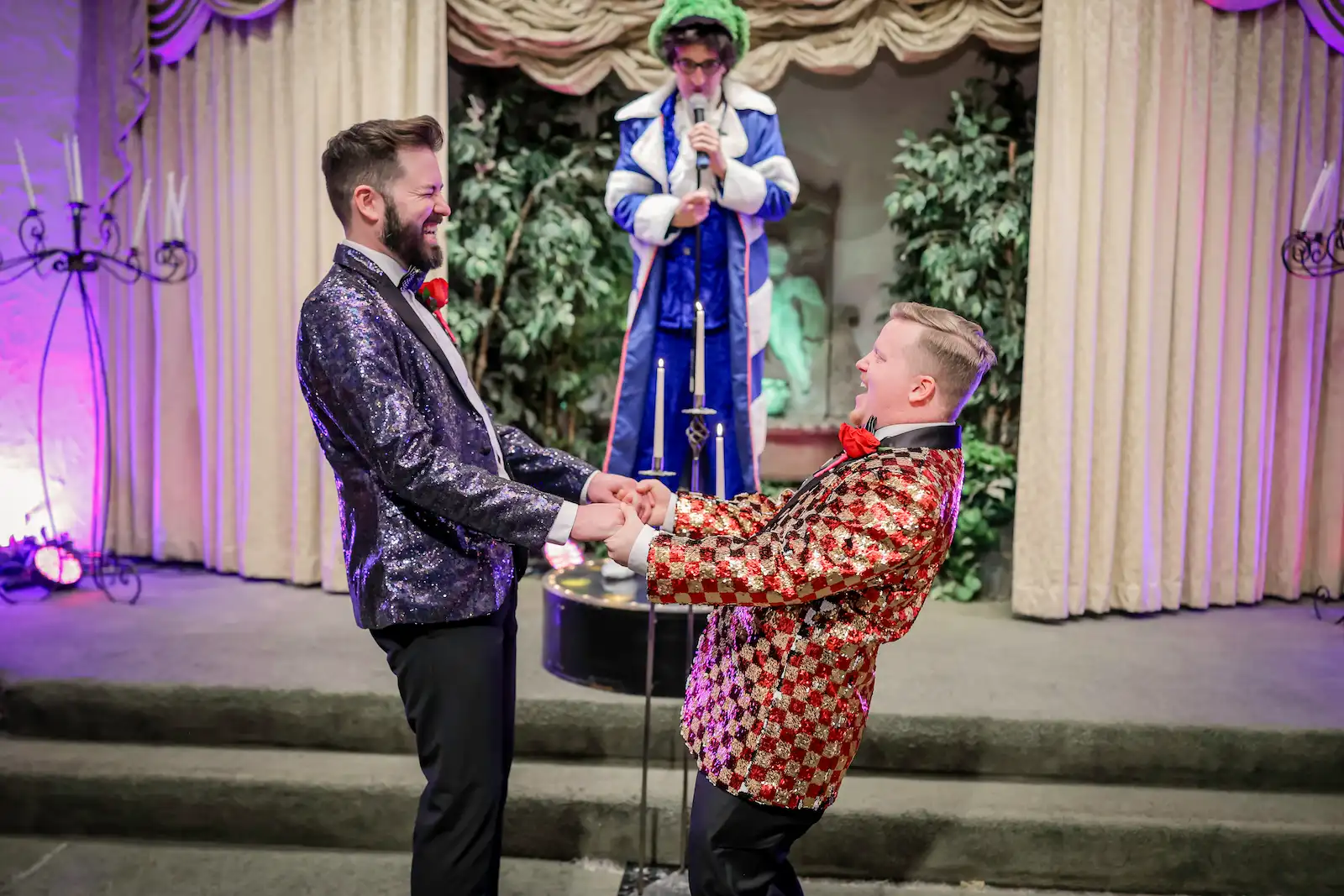 Two grooms in sequin blazers holding hands and laughing with Austin Powers impersonator officiating at a Las Vegas wedding chapel