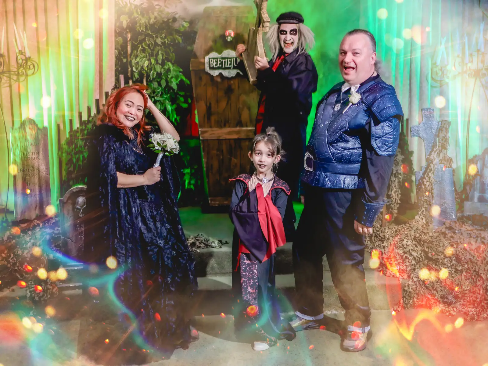 Family in Halloween costumes posing with Beetlejuice impersonator by coffin on graveyard stage with sparkle effects at Viva Las Vegas themed wedding