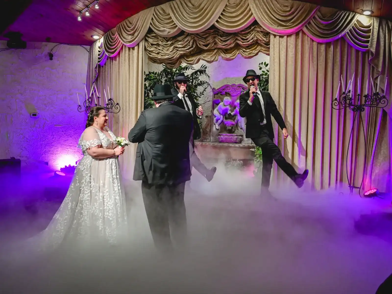 Blues Brothers impersonators in black suits, hats, and sunglasses performing and dancing in stage fog as bride laughs at Las Vegas themed wedding