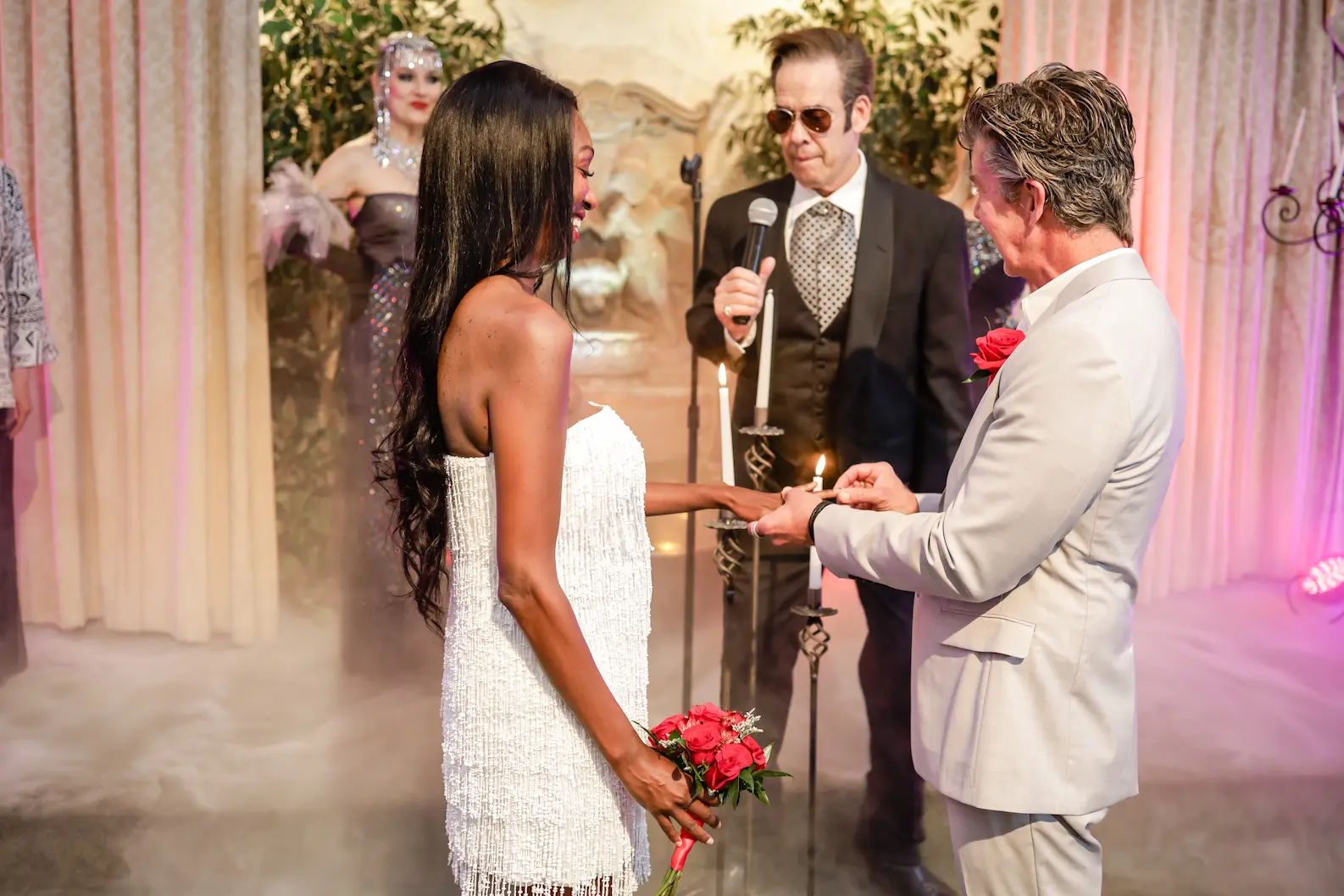Couple exchanging rings as James Bond impersonator in sunglasses and tuxedo officiates with showgirl on stage at Las Vegas Bond themed wedding