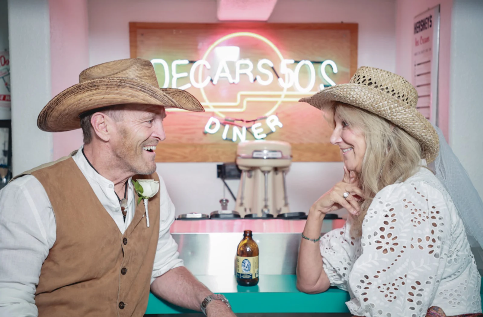 Vintage Las Vegas wedding couple wearing cowboy hats smiling at each other inside retro diner with neon sign backdrop