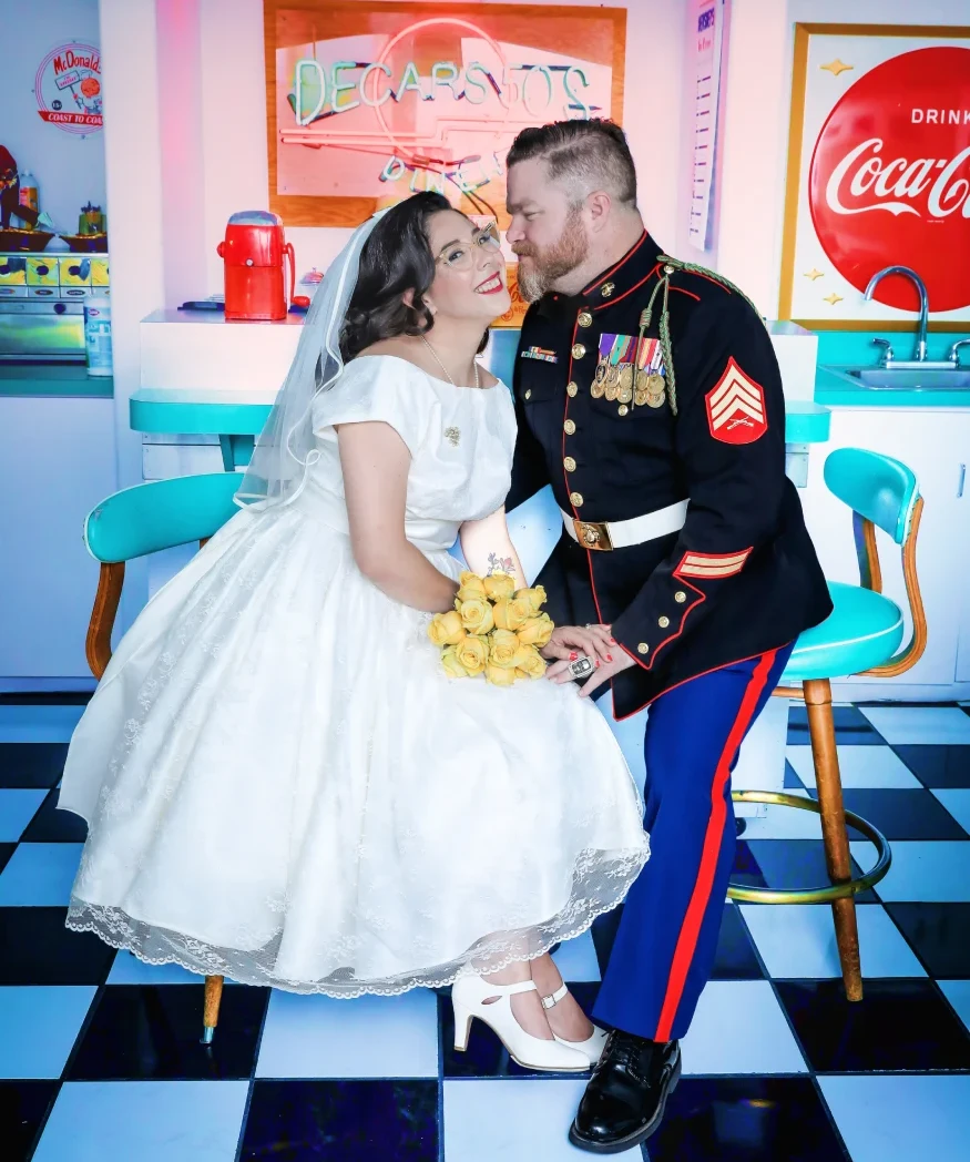 Retro Las Vegas diner wedding photo with bride in vintage dress and groom in military uniform sharing romantic moment beneath neon sign