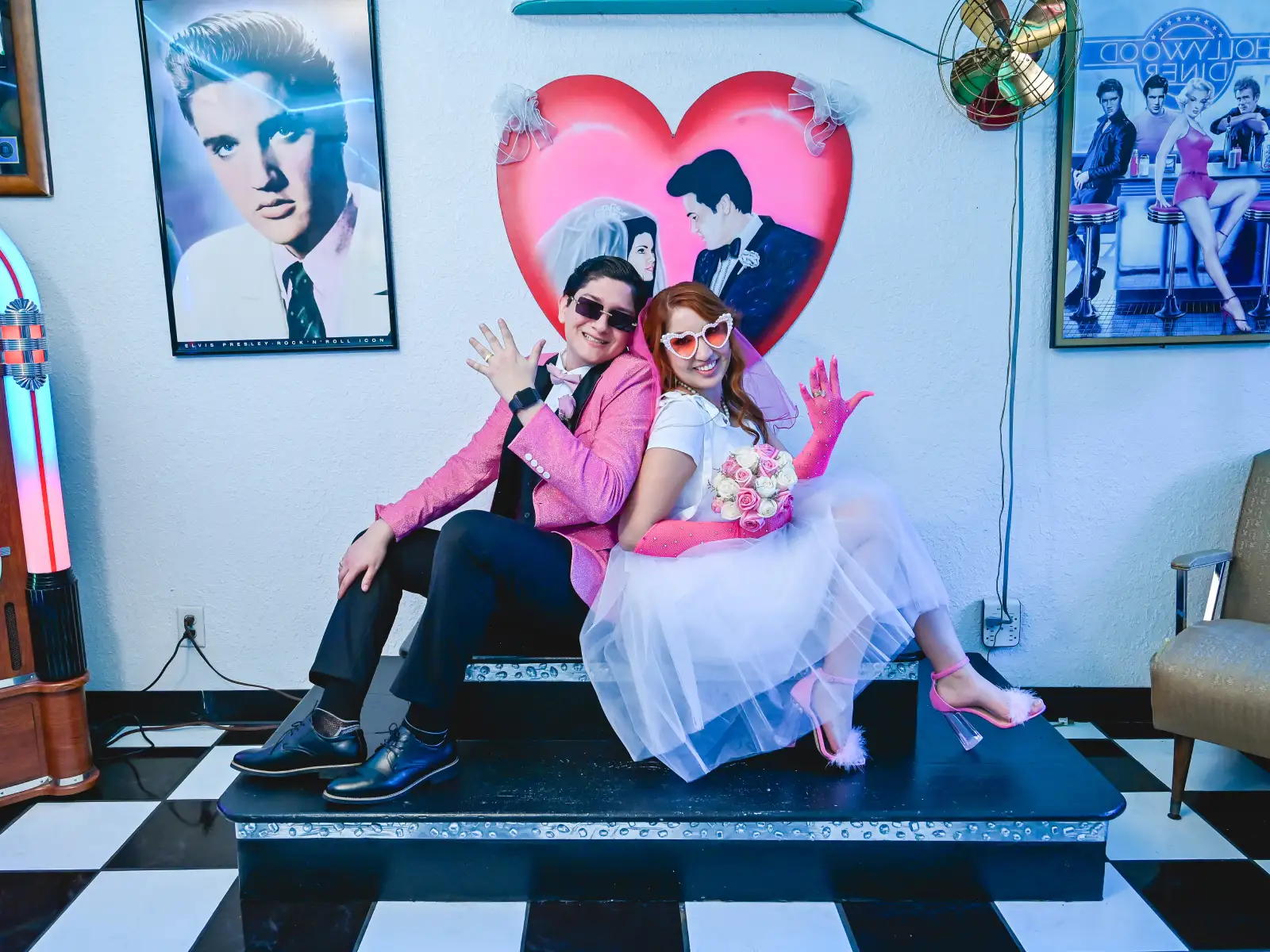 Fun retro Las Vegas wedding couple posing back to back in pink outfits inside rock-and-roll themed diner chapel with Elvis decor and heart backdrop