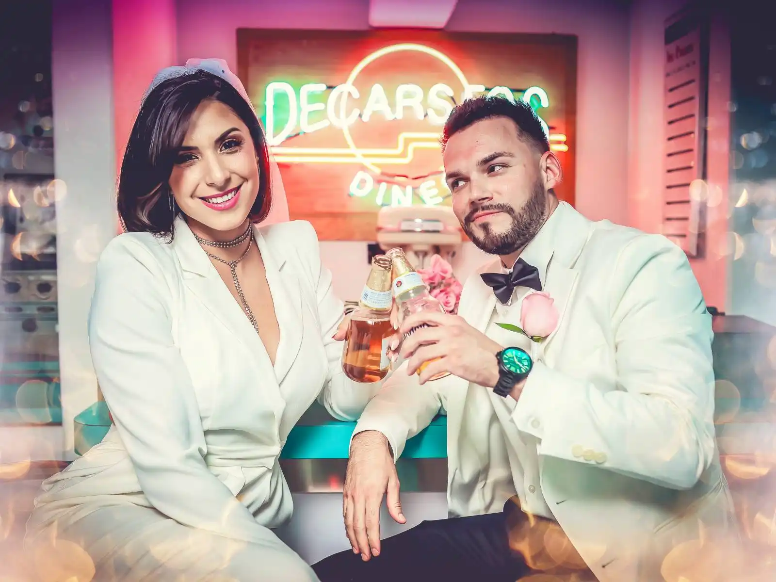 Couple in matching white suits toasting with beer bottles at a retro Vegas-style neon diner booth with pink roses and vintage decor