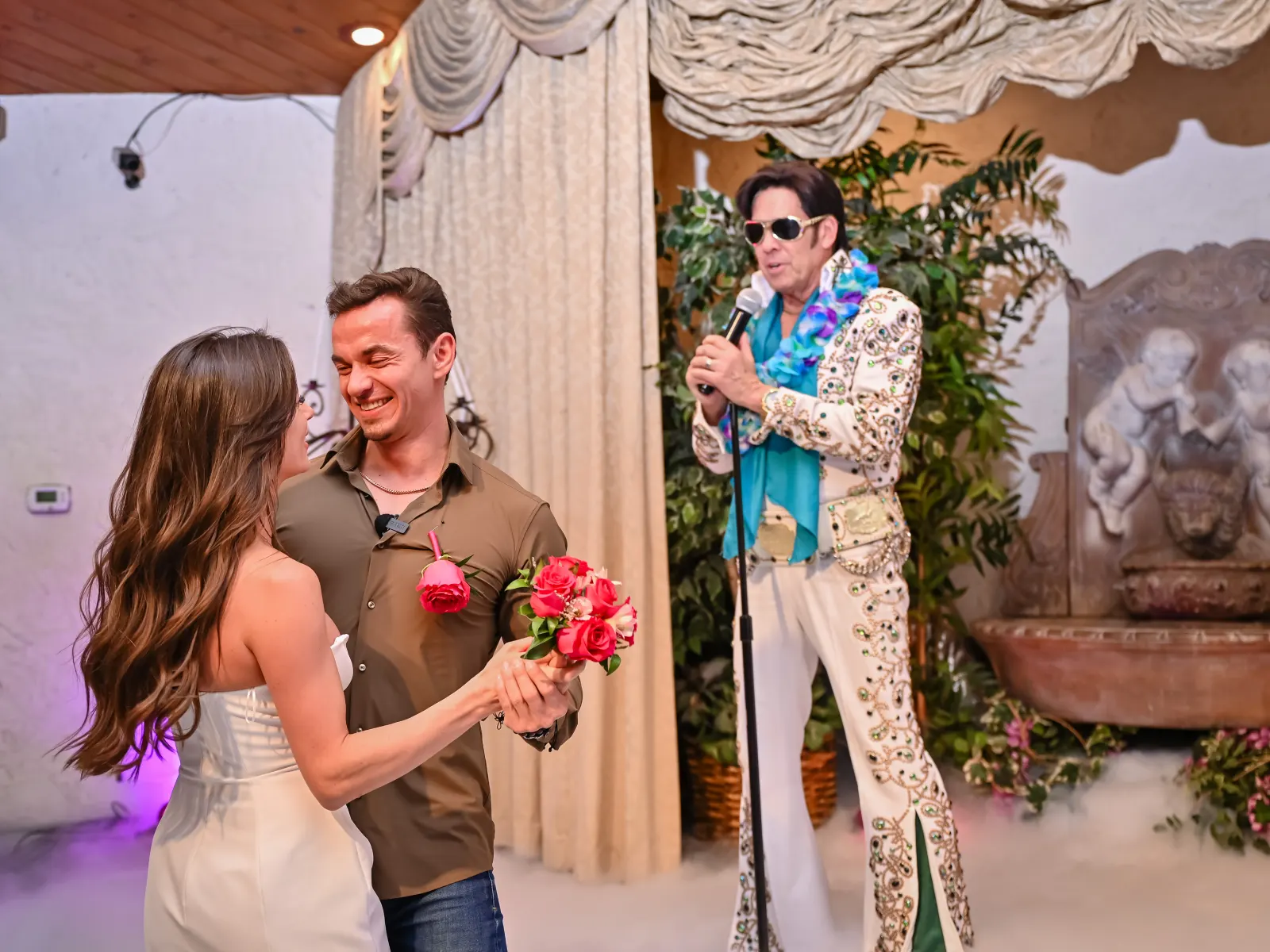 Elvis impersonator performing romantic song for smiling couple holding red rose bouquet during fun chapel wedding ceremony
