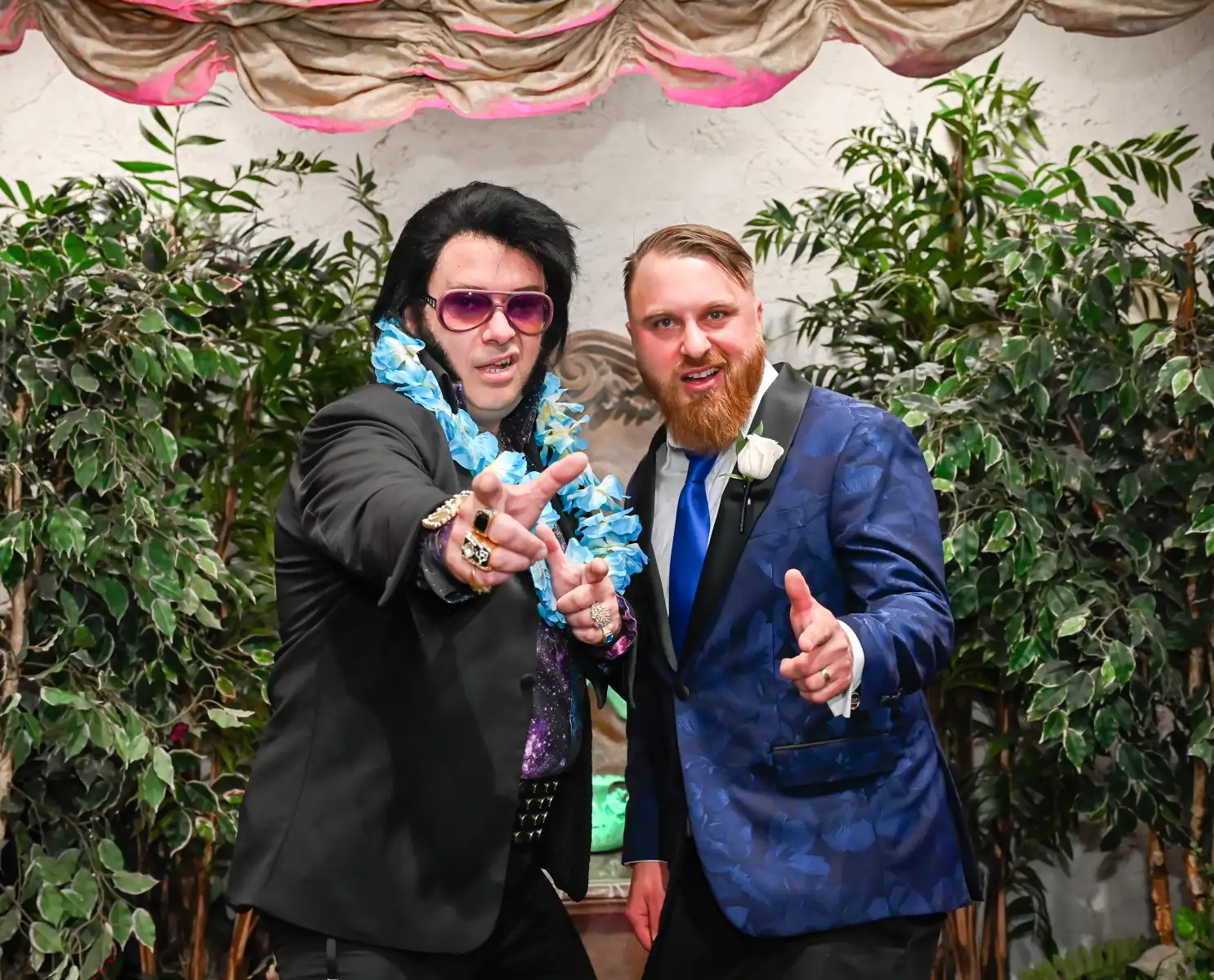 Elvis impersonator in stylish black suit and purple sunglasses posing playfully with smiling groom in blue patterned tuxedo during lively chapel wedding photo moment