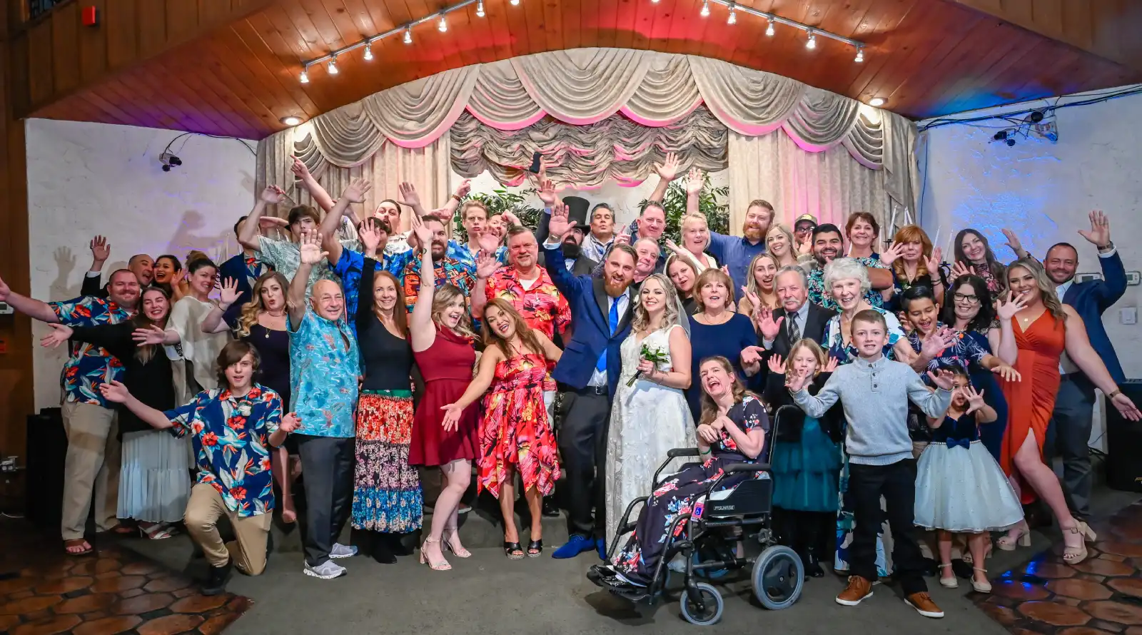 Large joyful wedding group photo with bride and groom surrounded by cheering family and friends posing together on decorated chapel stage under warm lights