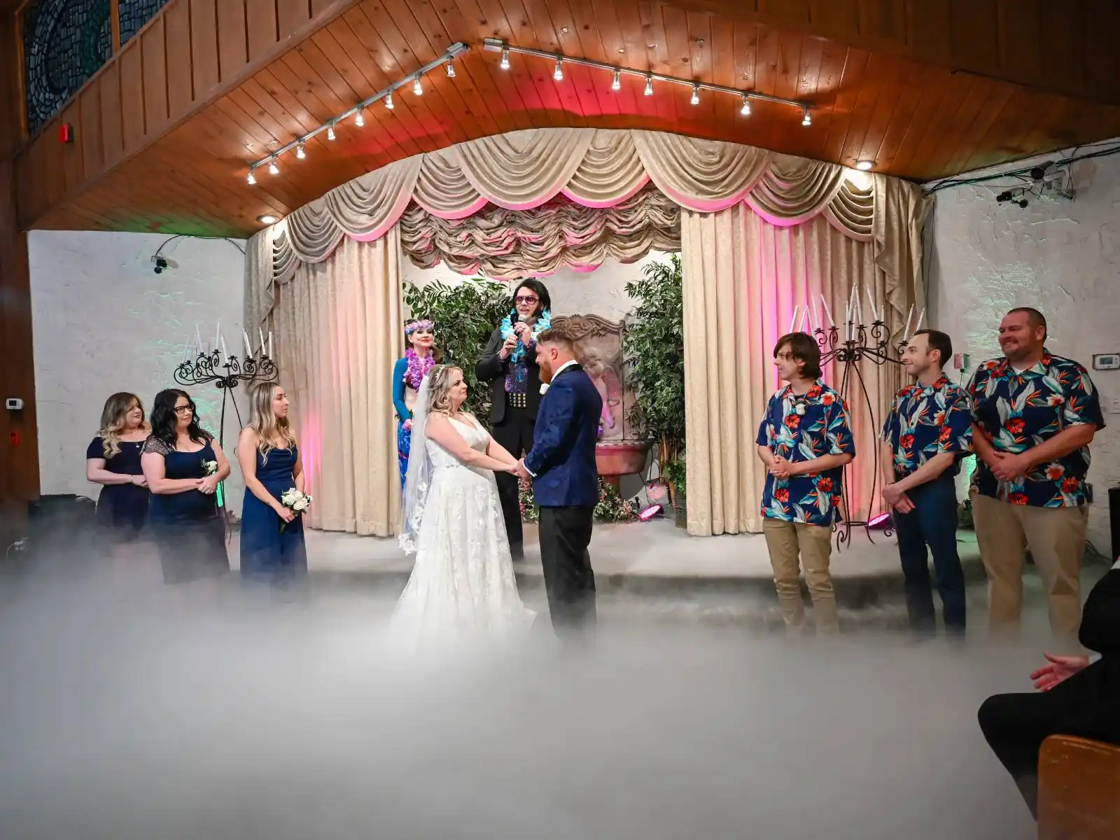 Couple exchanging vows with fog machine effects and Elvis officiant at a retro Las Vegas wedding chapel ceremony with bridal party