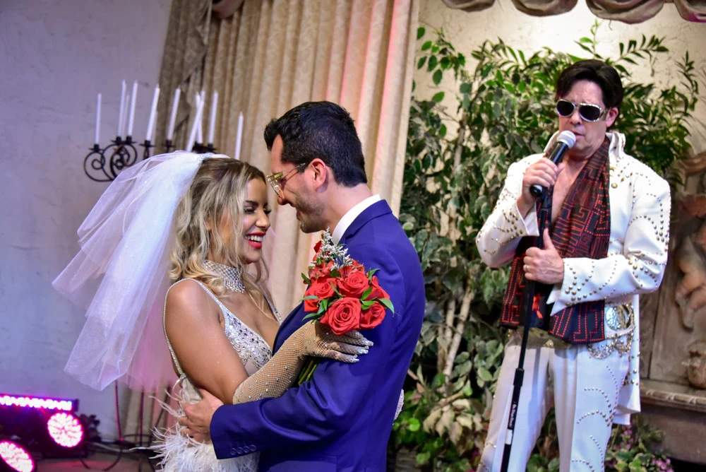 Romantic Las Vegas chapel wedding first dance with elegant bride and groom holding red roses while Elvis impersonator performs live nearby