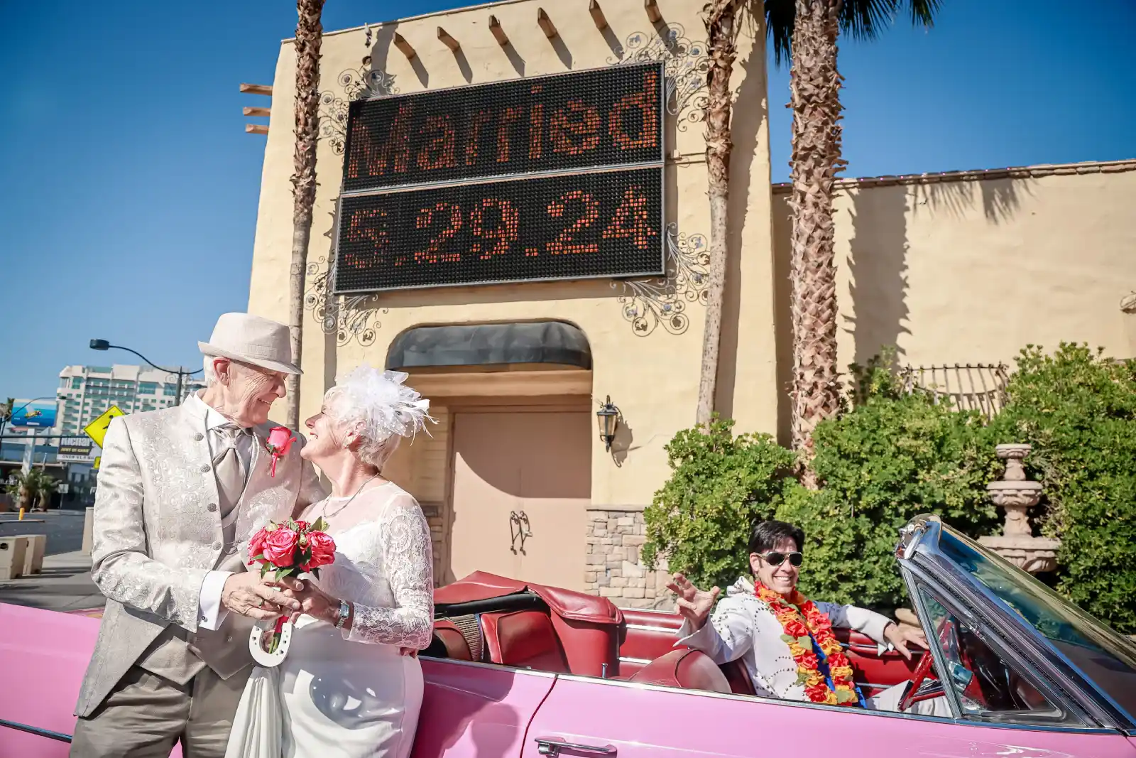 Elderly wedding couple holding bouquet beside pink Cadillac outside Las Vegas chapel sign