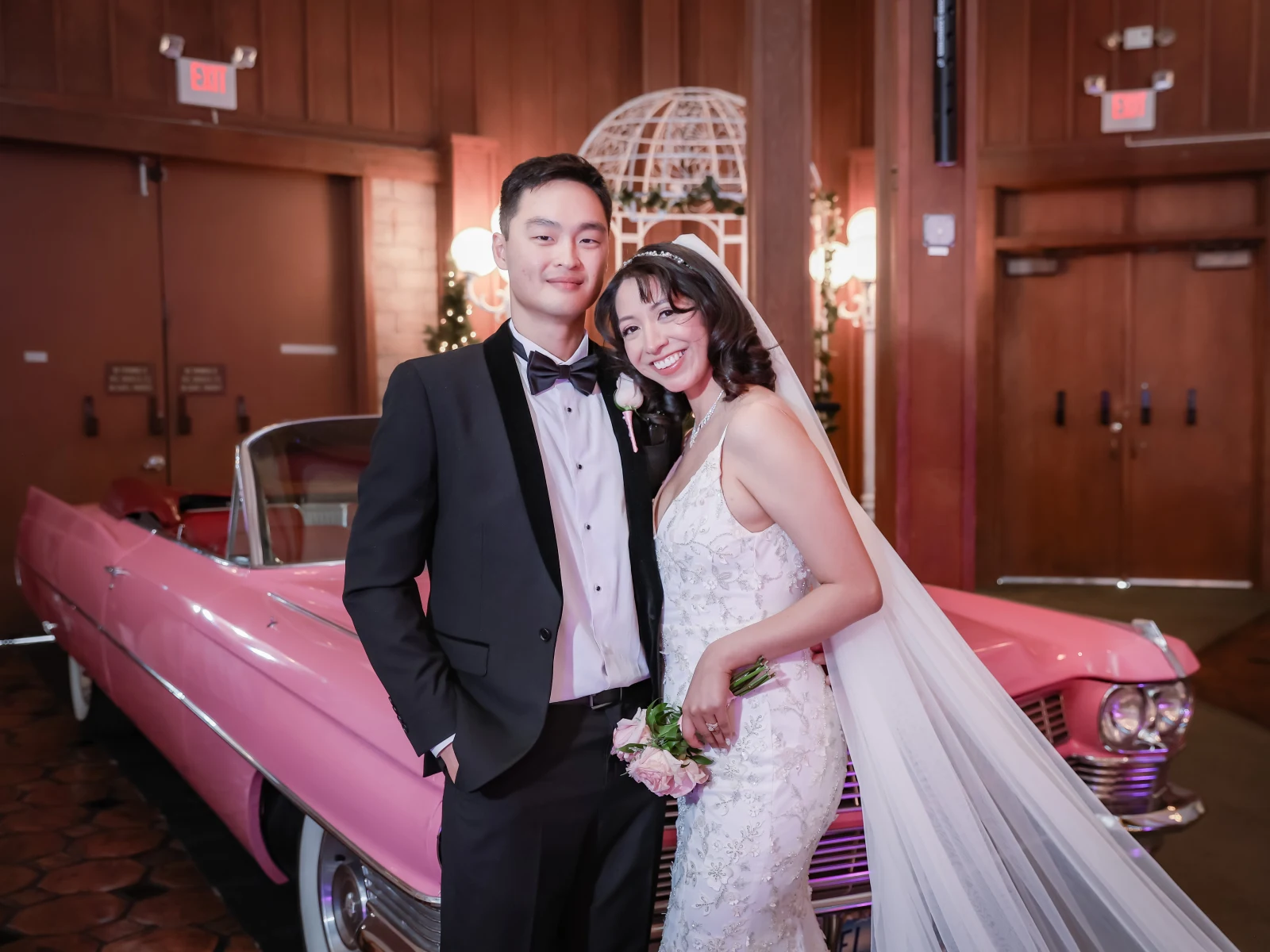 Bride and groom posing in formal wedding attire in front of pink vintage Cadillac indoors