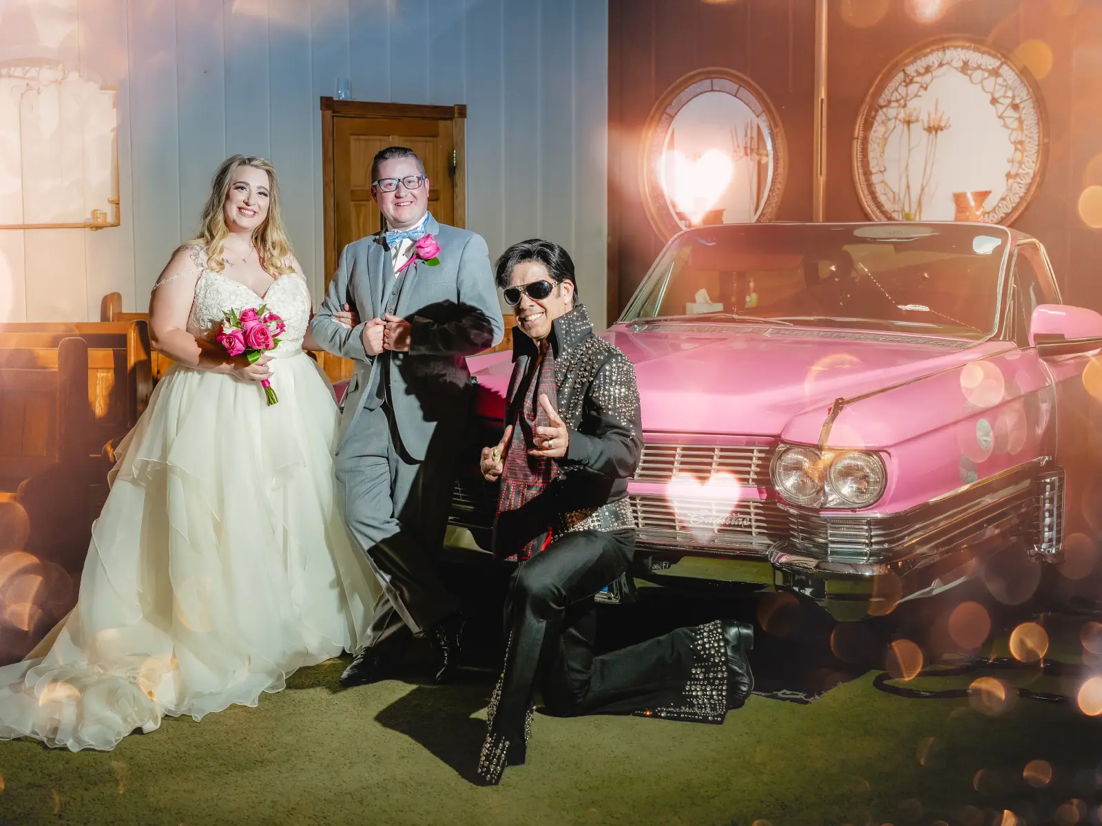Bride and groom posing beside pink vintage Cadillac with Elvis performer indoors