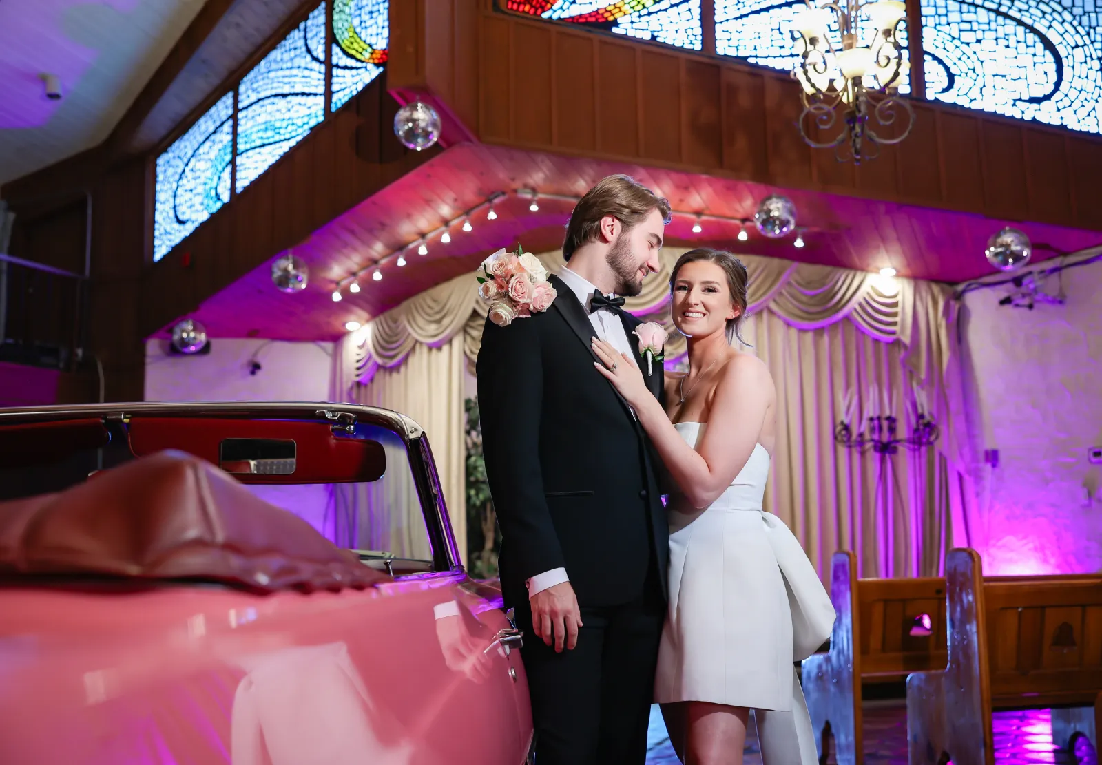 Bride touching groom’s lapel beside pink vintage convertible inside chapel with stained glass