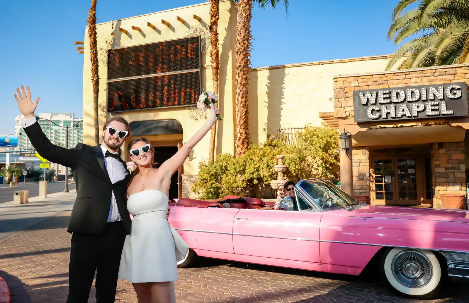 lBride and groom wearing heart-shaped sunglasses raising arms beside pink vintage convertible outside wedding chapel sign