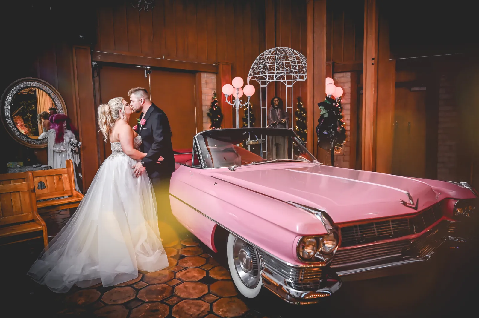 Bride and groom kissing beside pink vintage Cadillac convertible inside wooden wedding chapel with warm lighting