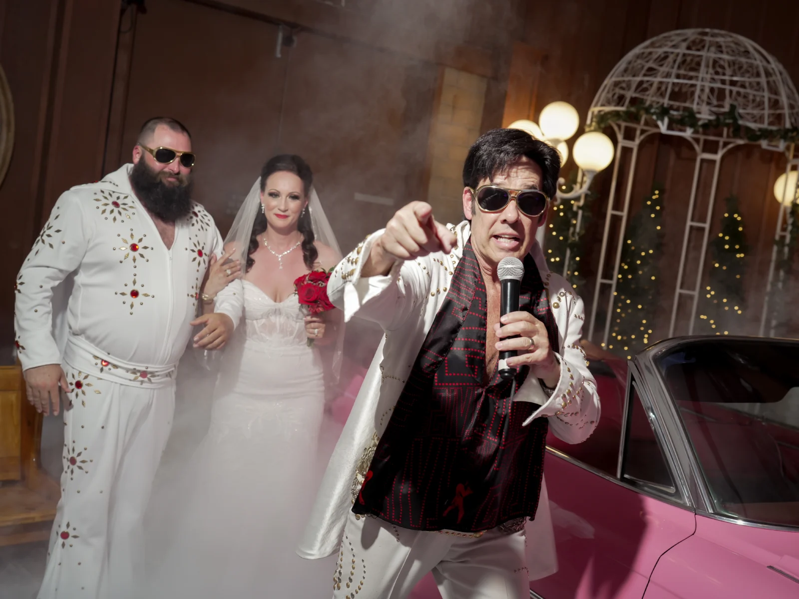 Elvis-style officiant pointing toward camera as newlyweds emerge through fog beside pink classic car in chapel entrance