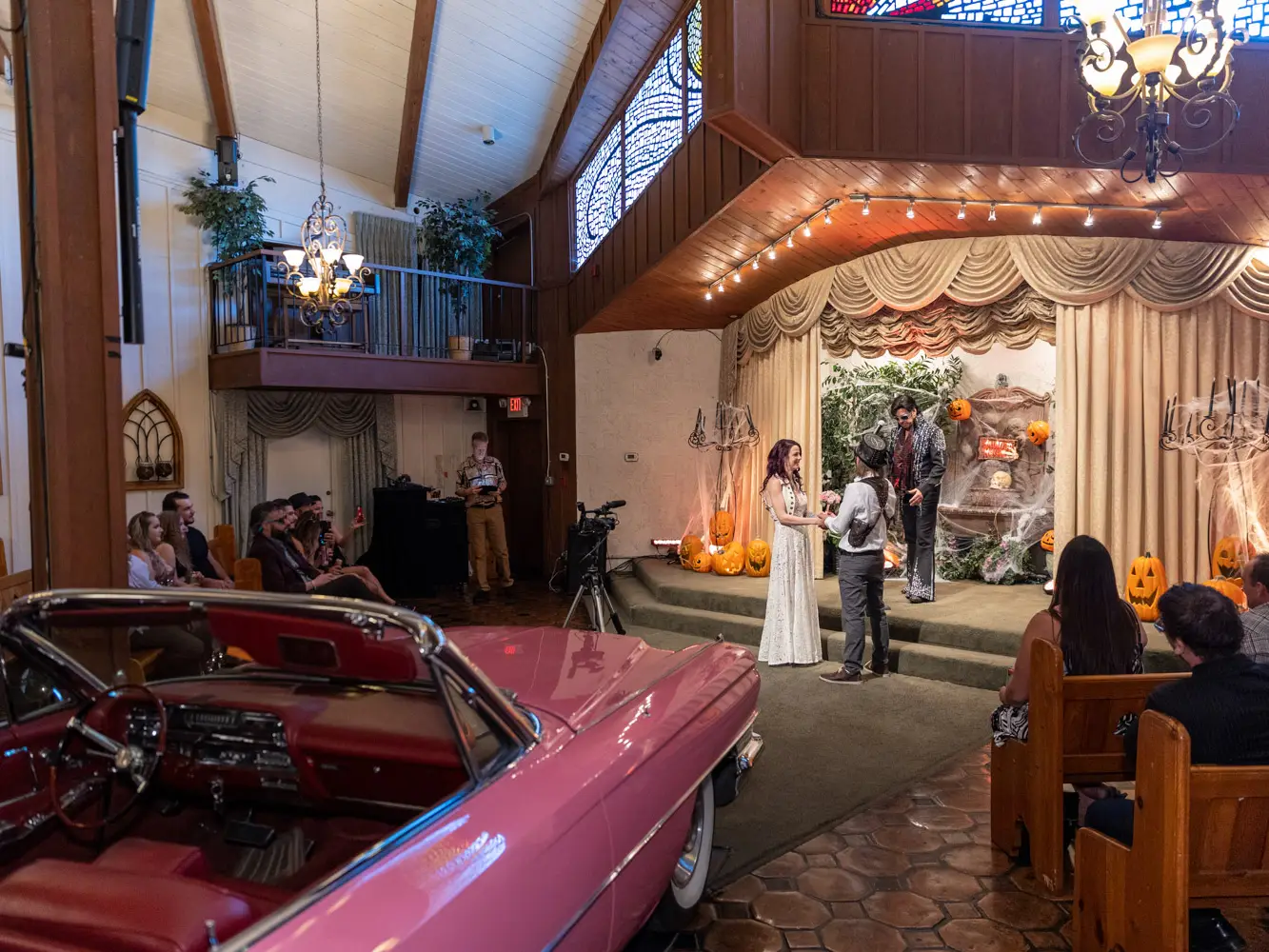 Wide chapel view with pink vintage car in foreground as couple stands on stage with Elvis officiant and seated guests watching
