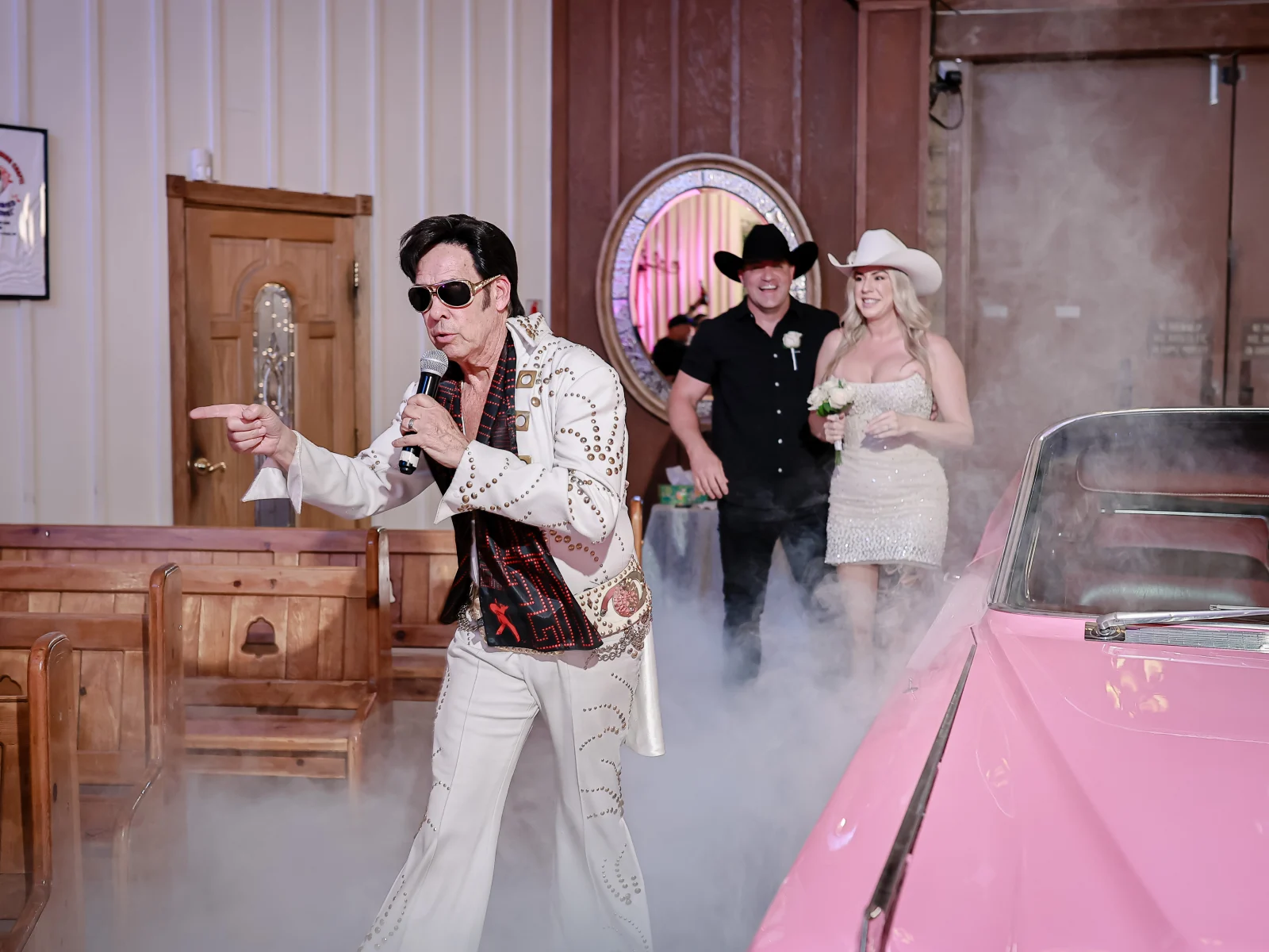 Elvis impersonator leading smiling cowboy-hat couple through chapel aisle beside pink vintage car and stage fog