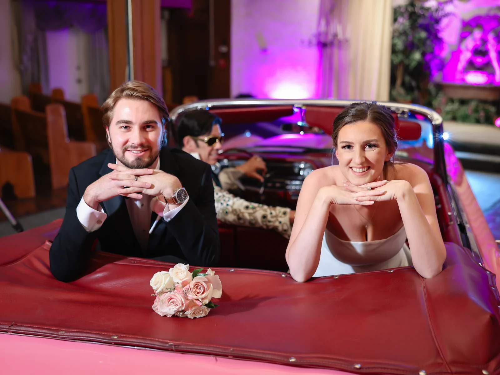Smiling newlywed couple leaning on pink convertible backseat with bouquet while Elvis impersonator drives inside chapel