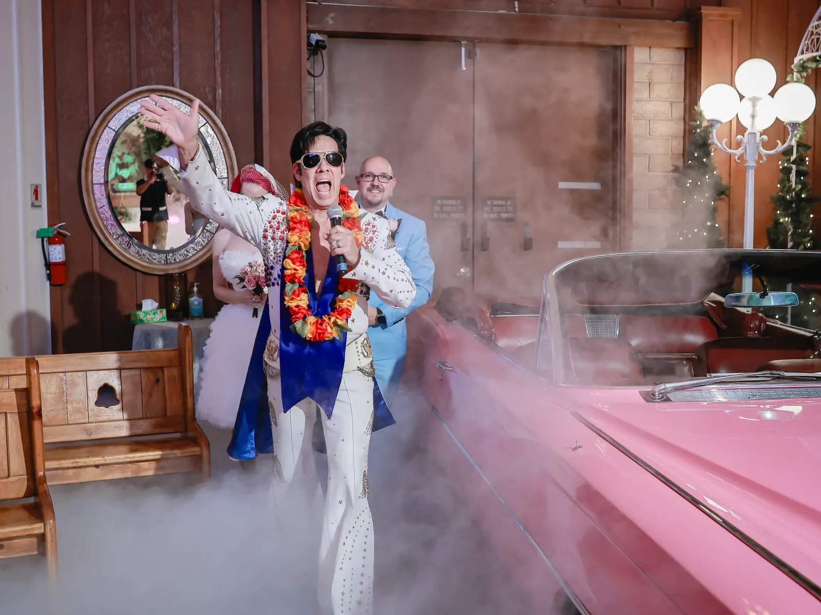 Elvis officiant singing beside pink convertible as newlyweds enter smoke-filled chapel aisle