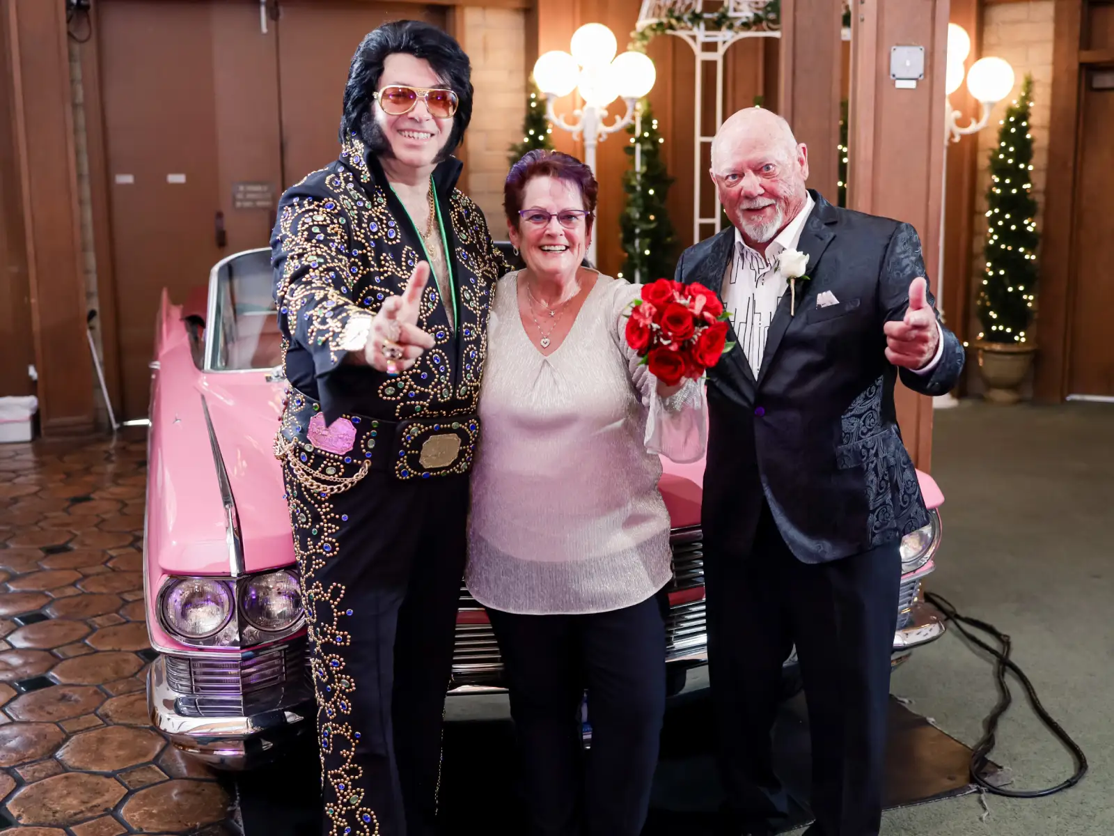 Elvis impersonator posing with smiling couple holding red rose bouquet in front of pink classic car