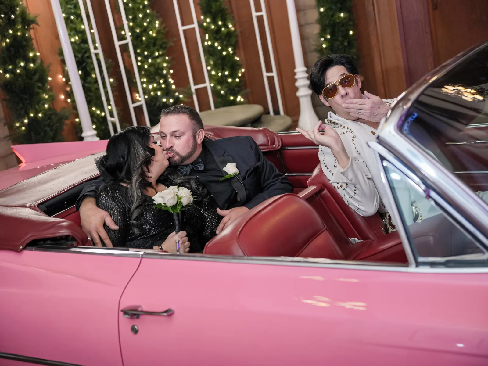 Bride and groom kissing in pink vintage convertible while Elvis impersonator blows a kiss at fun wedding chapel ceremony