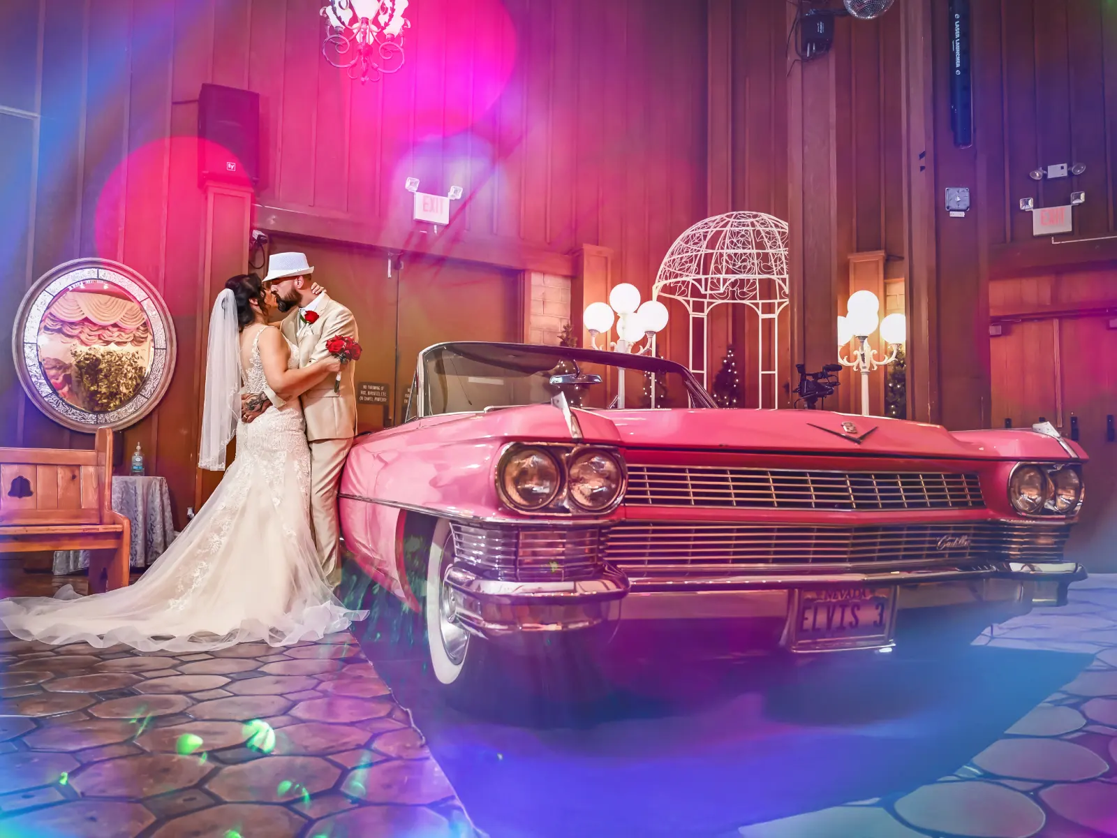 Bride and groom kissing beside pink vintage Cadillac convertible with colorful lens flare lighting inside fun wedding chapel