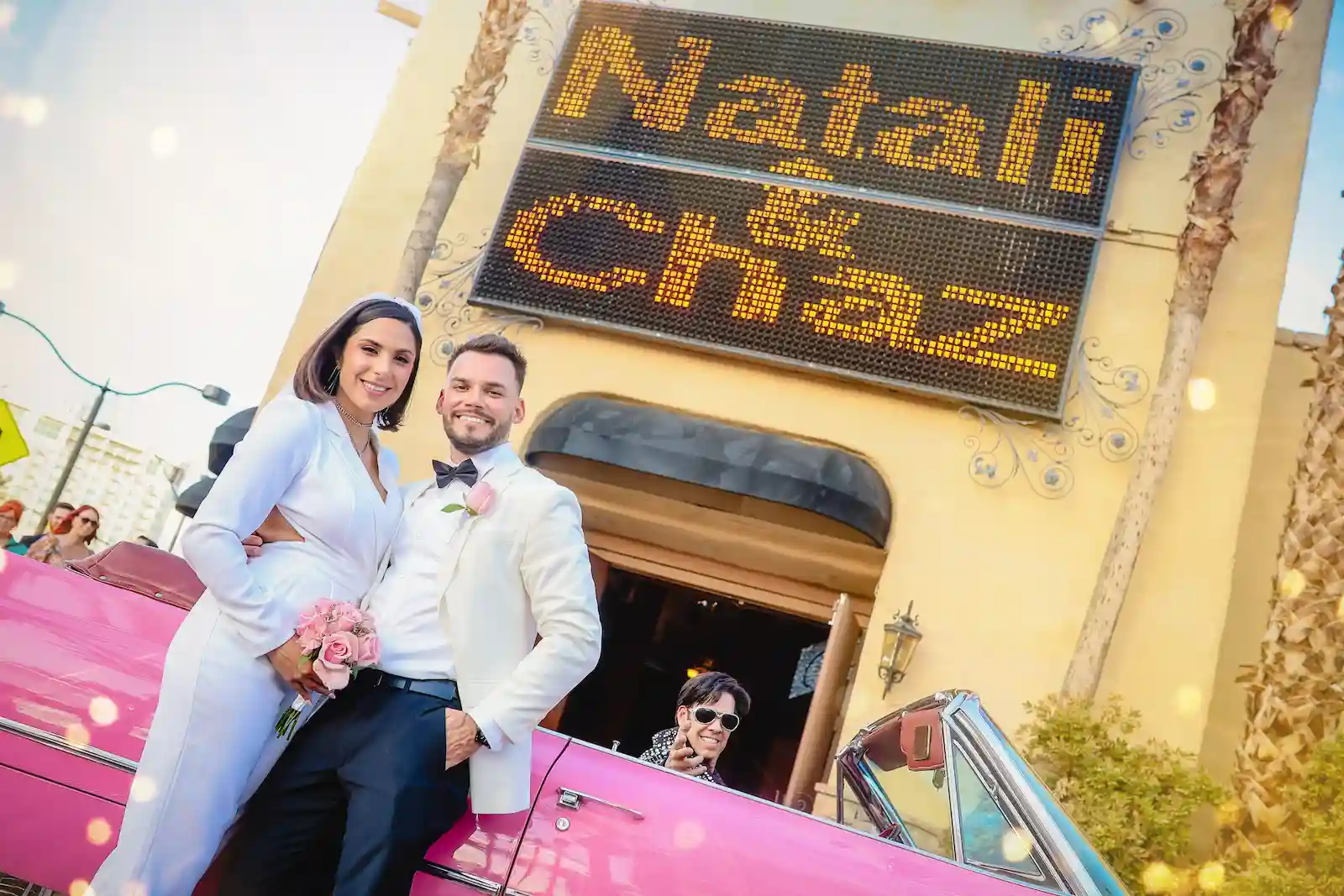 Newlywed couple posing with pink Cadillac and Elvis impersonator outside a retro Las Vegas wedding chapel with marquee sign