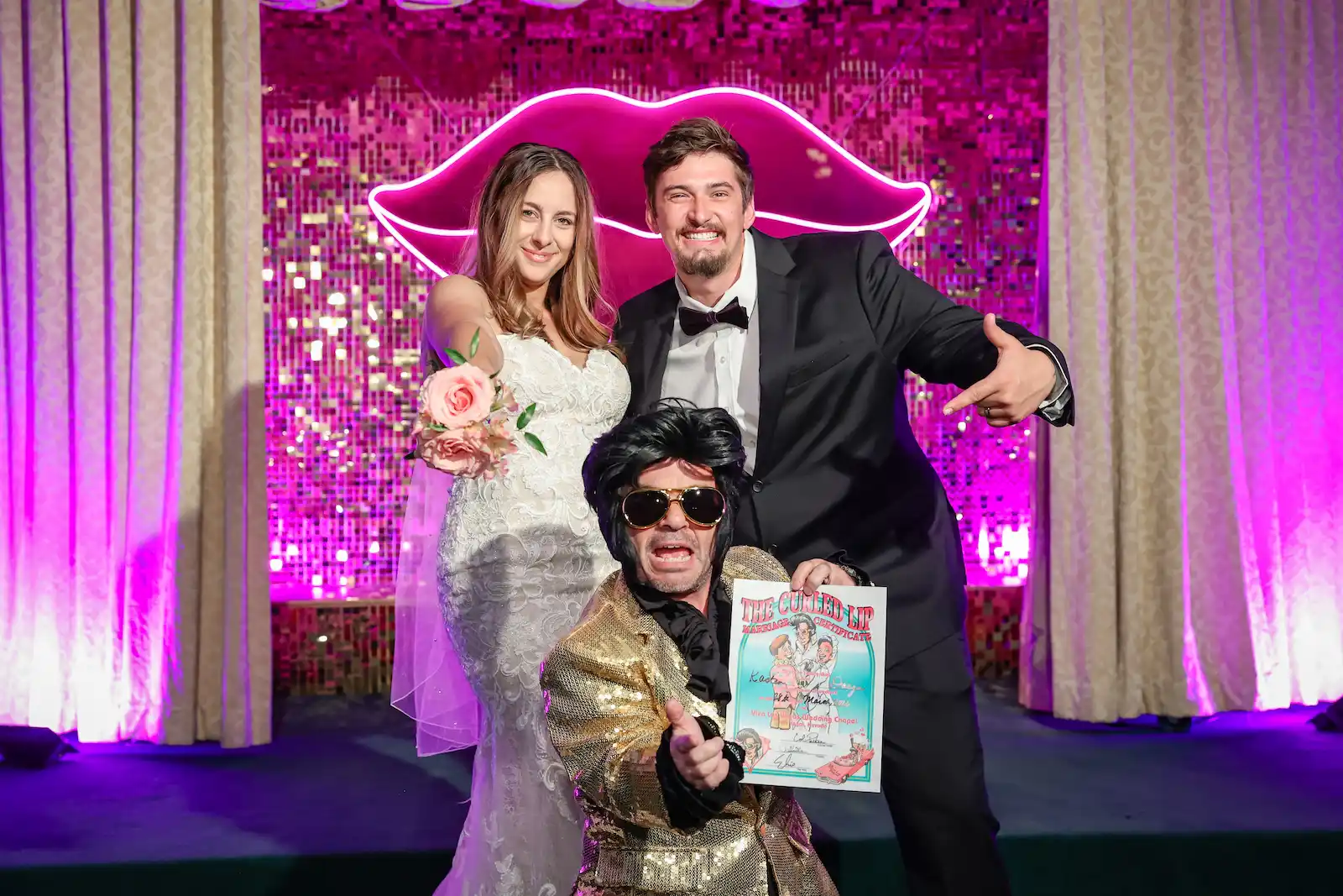 Bride and groom posing with Little Elvis impersonator holding a marriage certificate in front of a neon lips sign at a Las Vegas wedding chapel