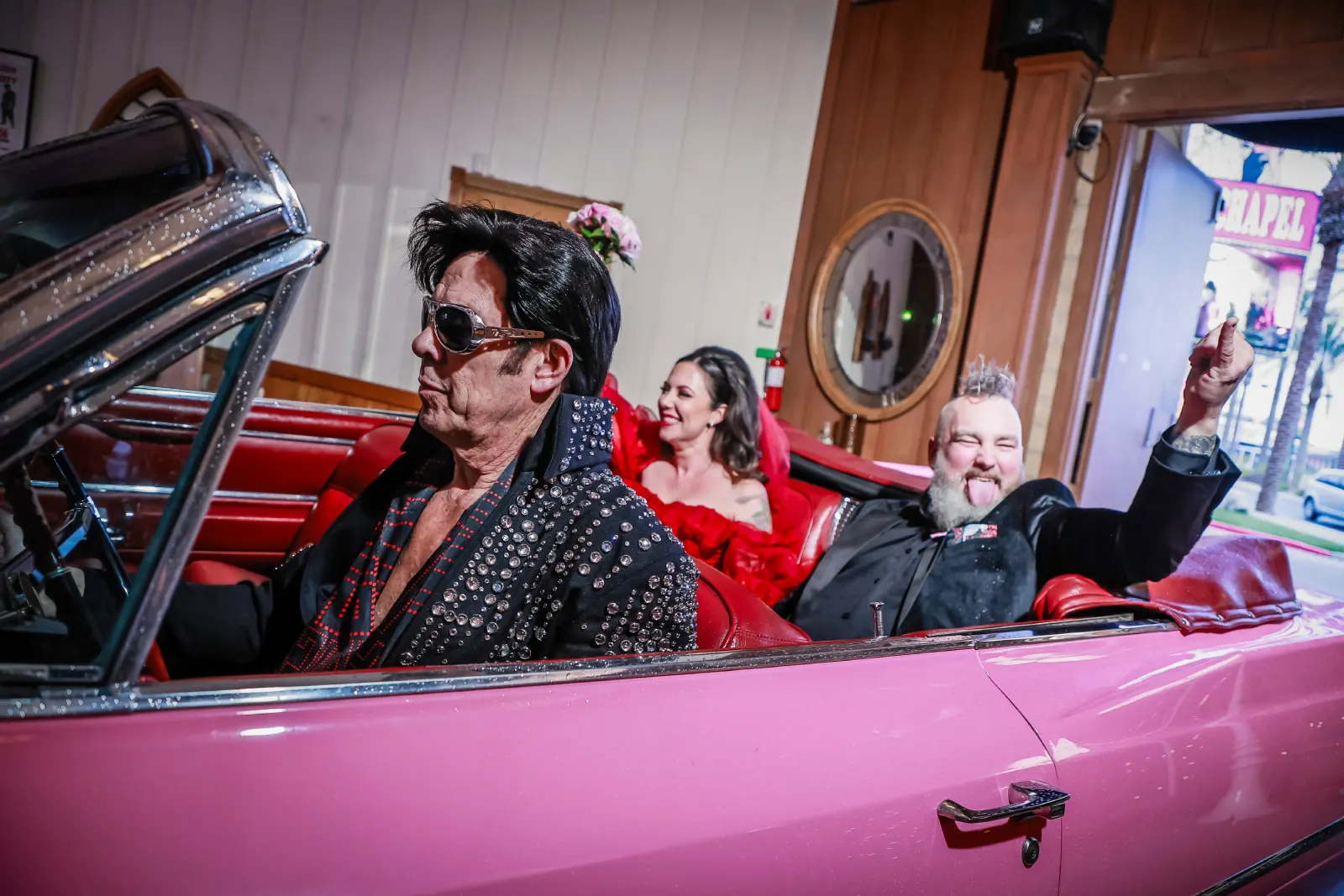 Elvis impersonator driving pink vintage convertible with laughing wedding guests in red and black outfits inside chapel entrance