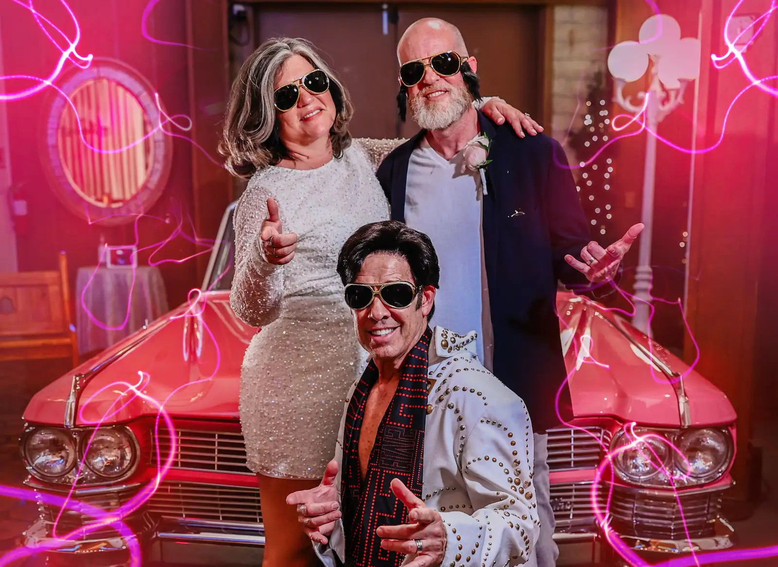 Bride, groom and Elvis impersonator posing with sunglasses in front of the pink Cadillac at Viva Las Vegas Wedding Chapel.