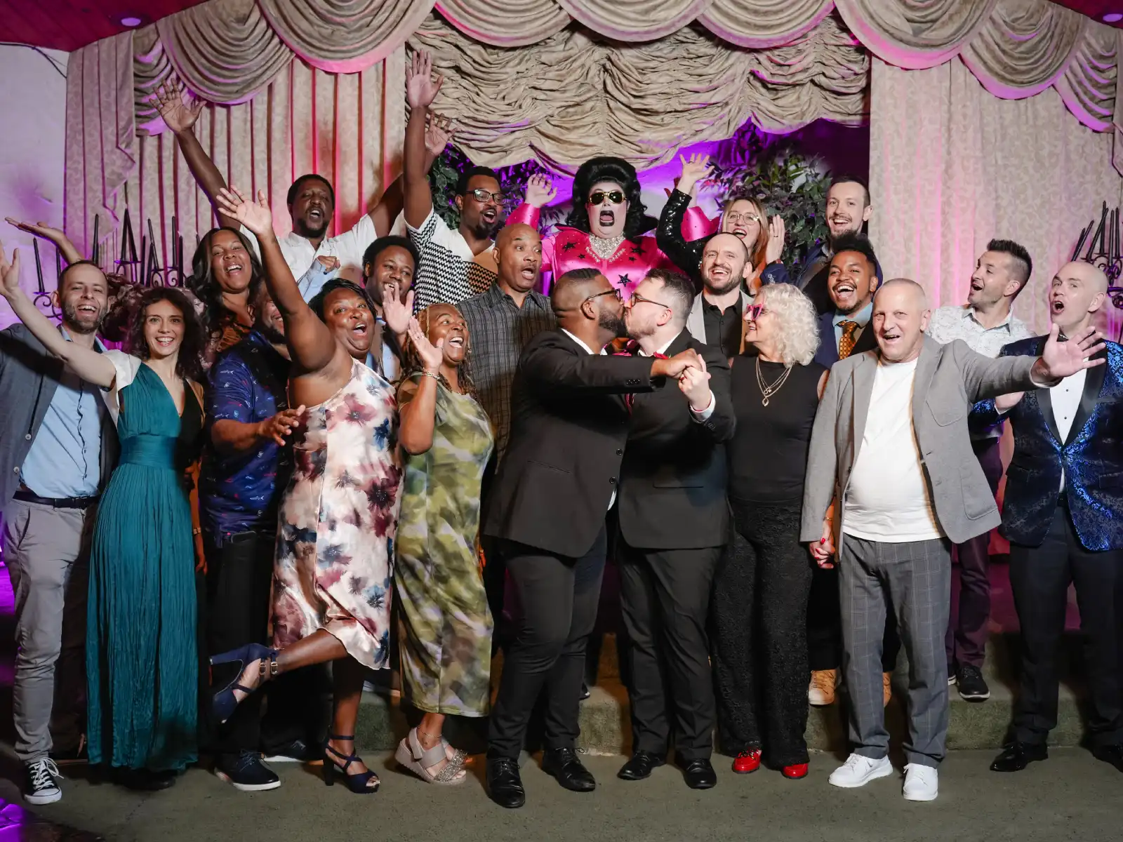 A large group of wedding guests posing for a photo on stage with the two grooms kissing in the center and a drag queen Elvis impersonator standing behind them.