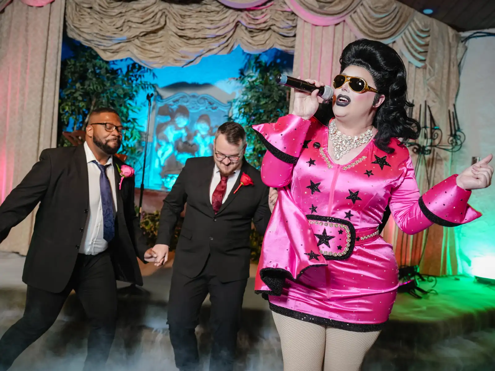 A drag queen Elvis Presley performer singing at a microphone on stage while the two grooms behind her dance during their wedding ceremony.
