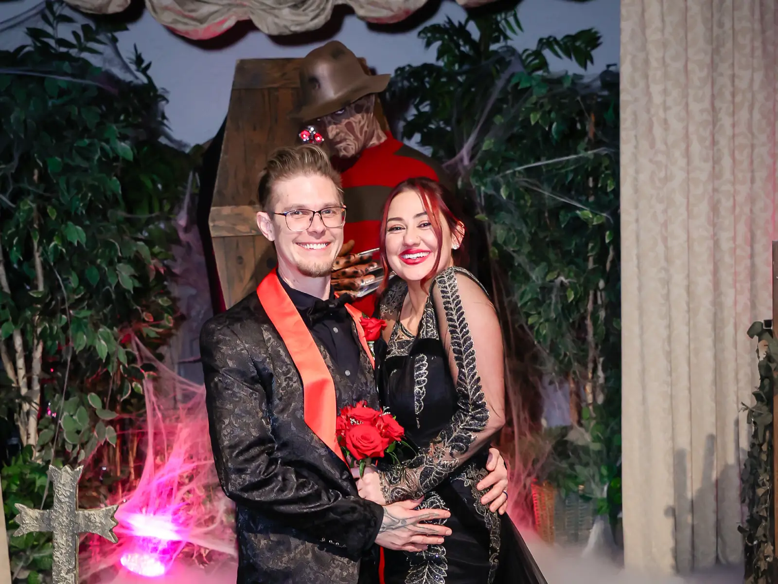 Newlywed couple smiling with red roses as Freddy Krueger impersonator lurks behind coffin at Viva Las Vegas horror themed wedding