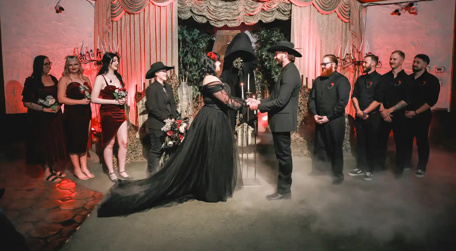 Bride in black gown exchanging vows with groom and wedding party bathed in red theatrical lighting with fog on gothic stage