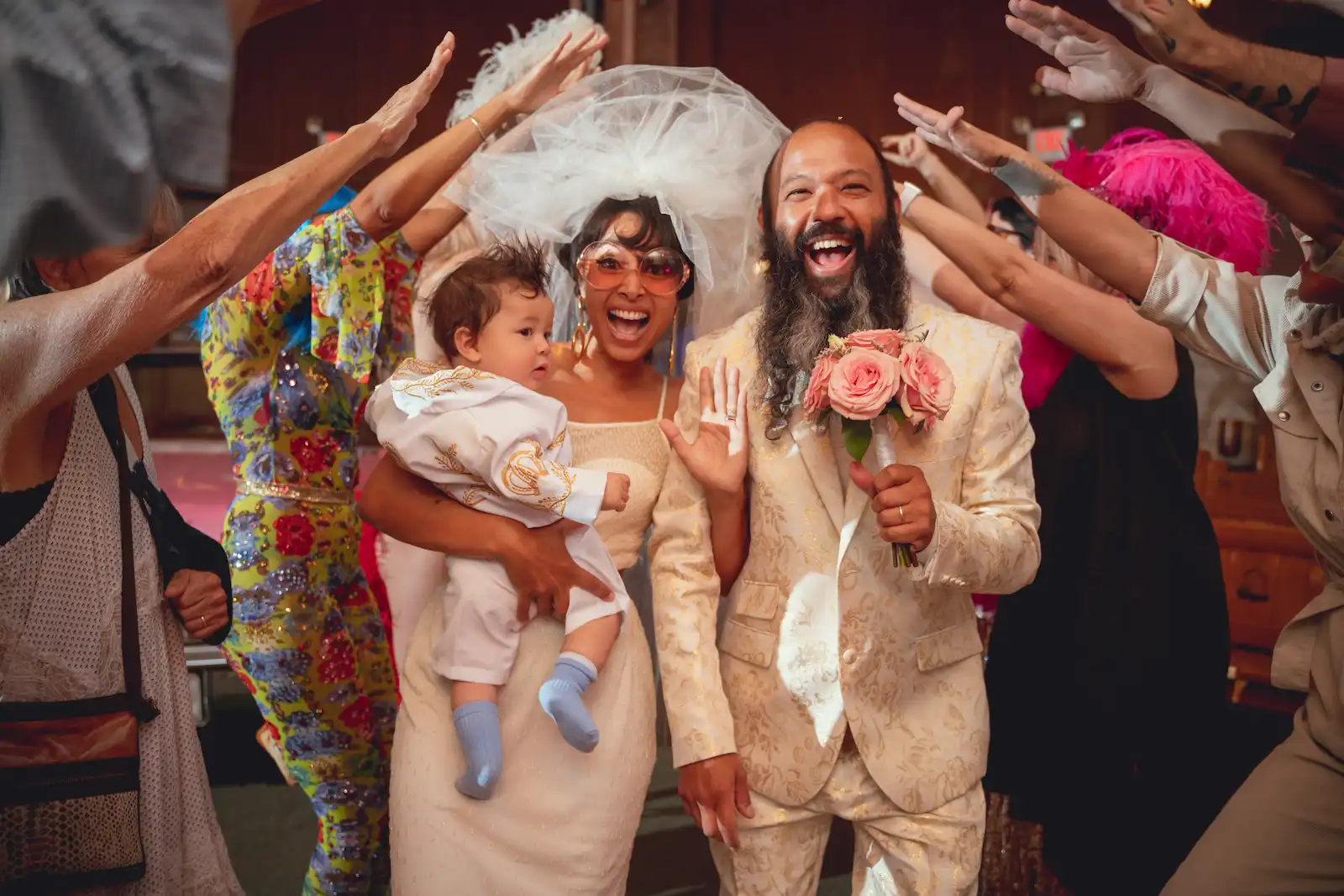 Bride holding baby and groom with pink bouquet laughing as guests raise hands in celebration at Las Vegas wedding chapel