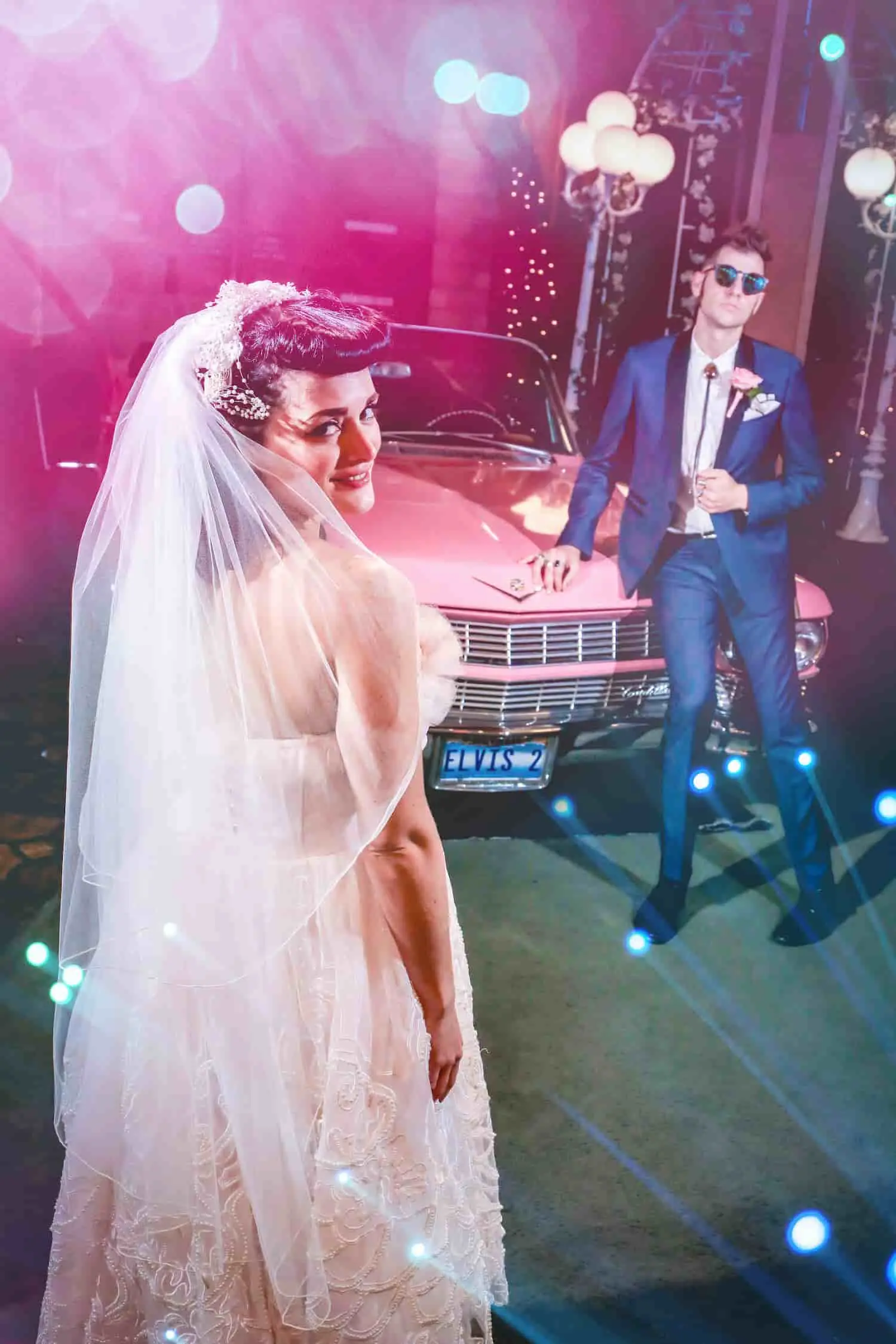 Bride in vintage veil and groom in blue suit posing with pink Cadillac under neon lights at Las Vegas wedding chapel
