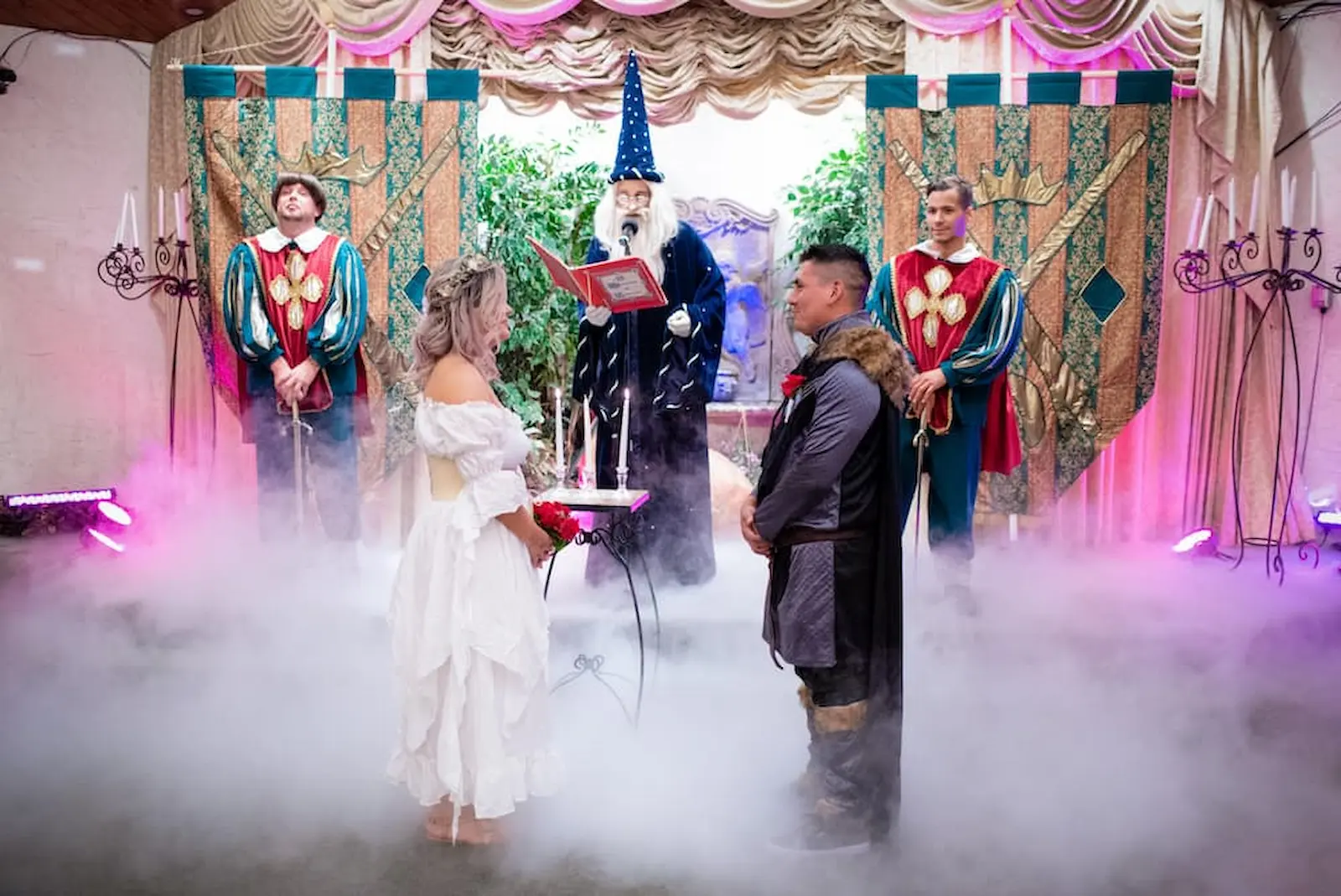 Couple exchanging vows with wizard officiant reading from spell book flanked by knights with banners and stage fog