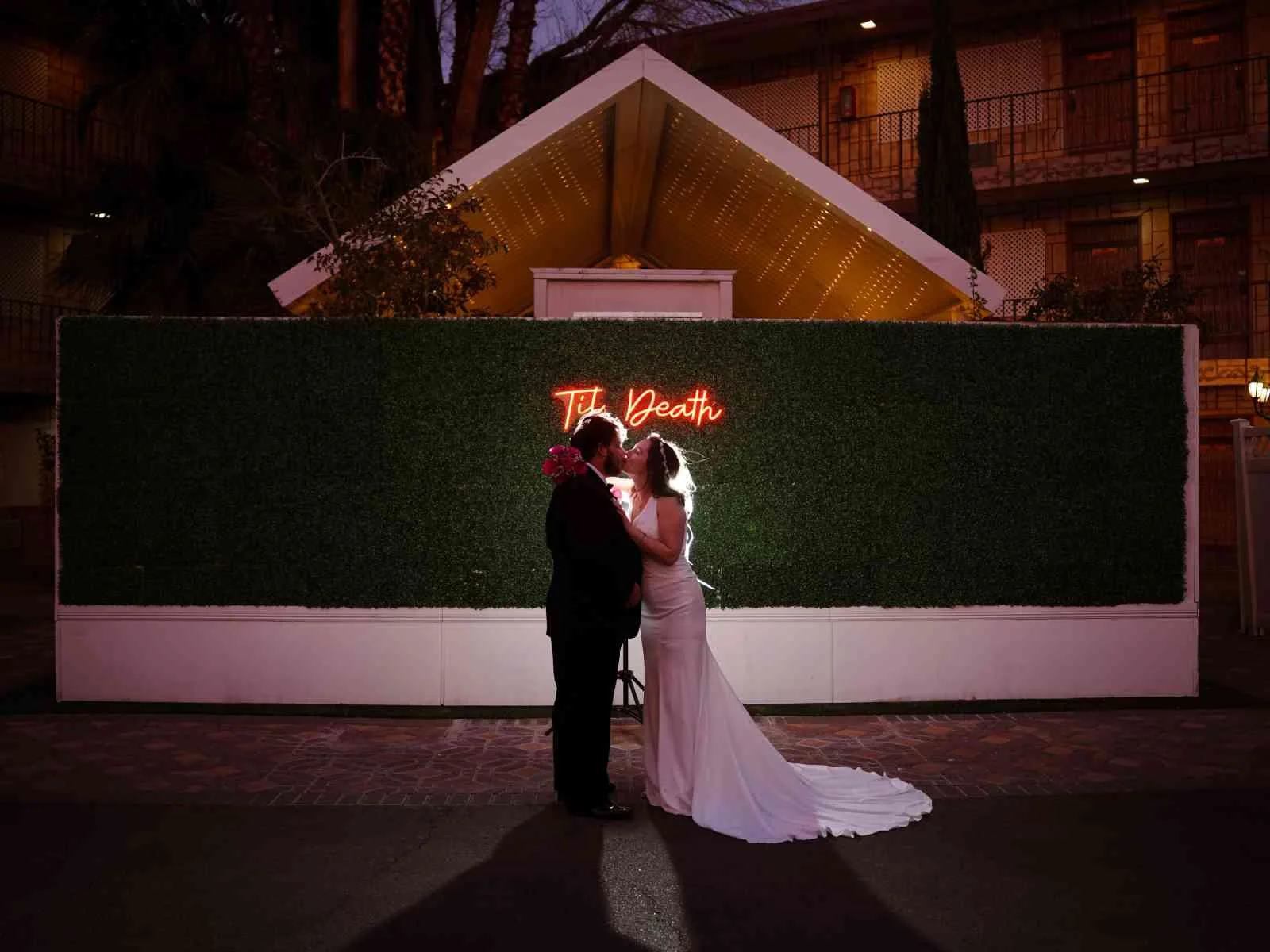 Couple kissing at night in front of til death neon sign on greenery wall at a Las Vegas wedding chapel