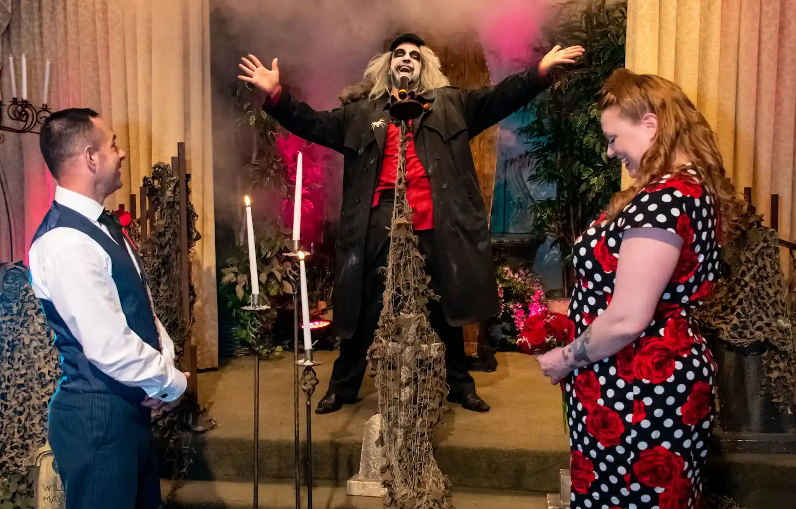 Beetlejuice impersonator with arms raised officiating wedding with candles and spooky decor on fog-filled stage