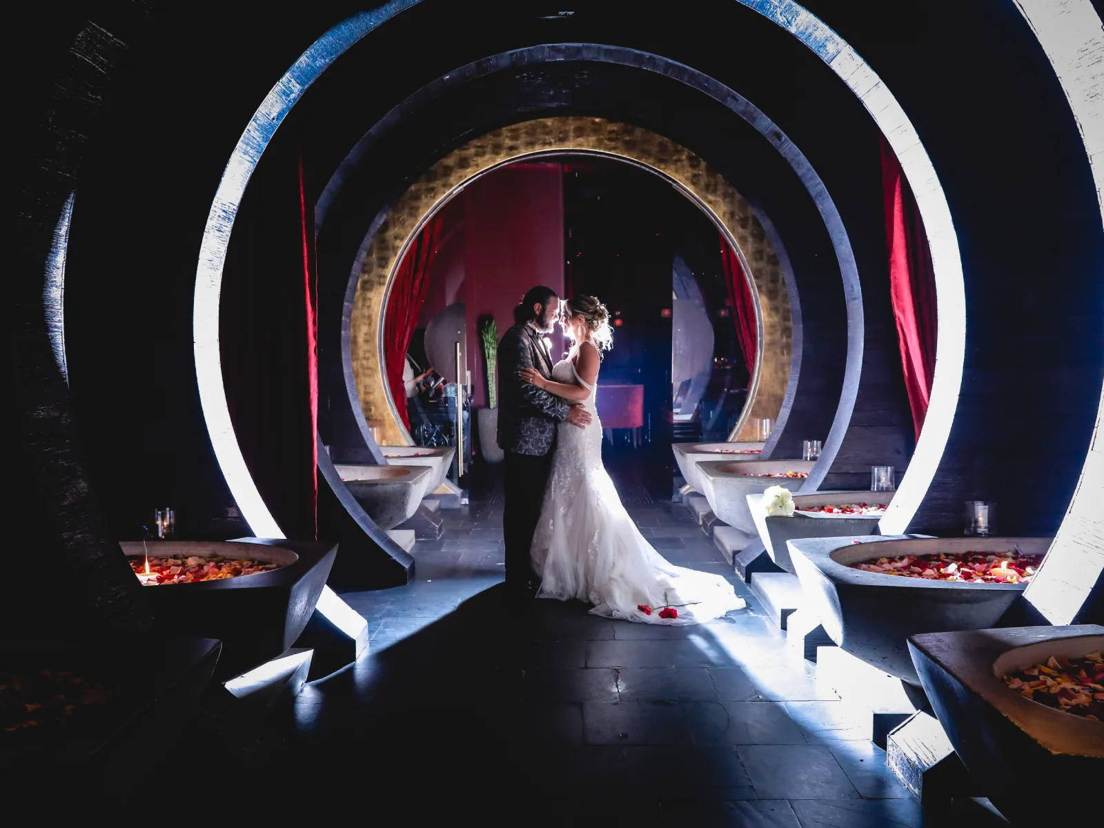 A bride and groom embracing in a dark, architecturally striking tunnel composed of glowing concentric circles and red curtains.