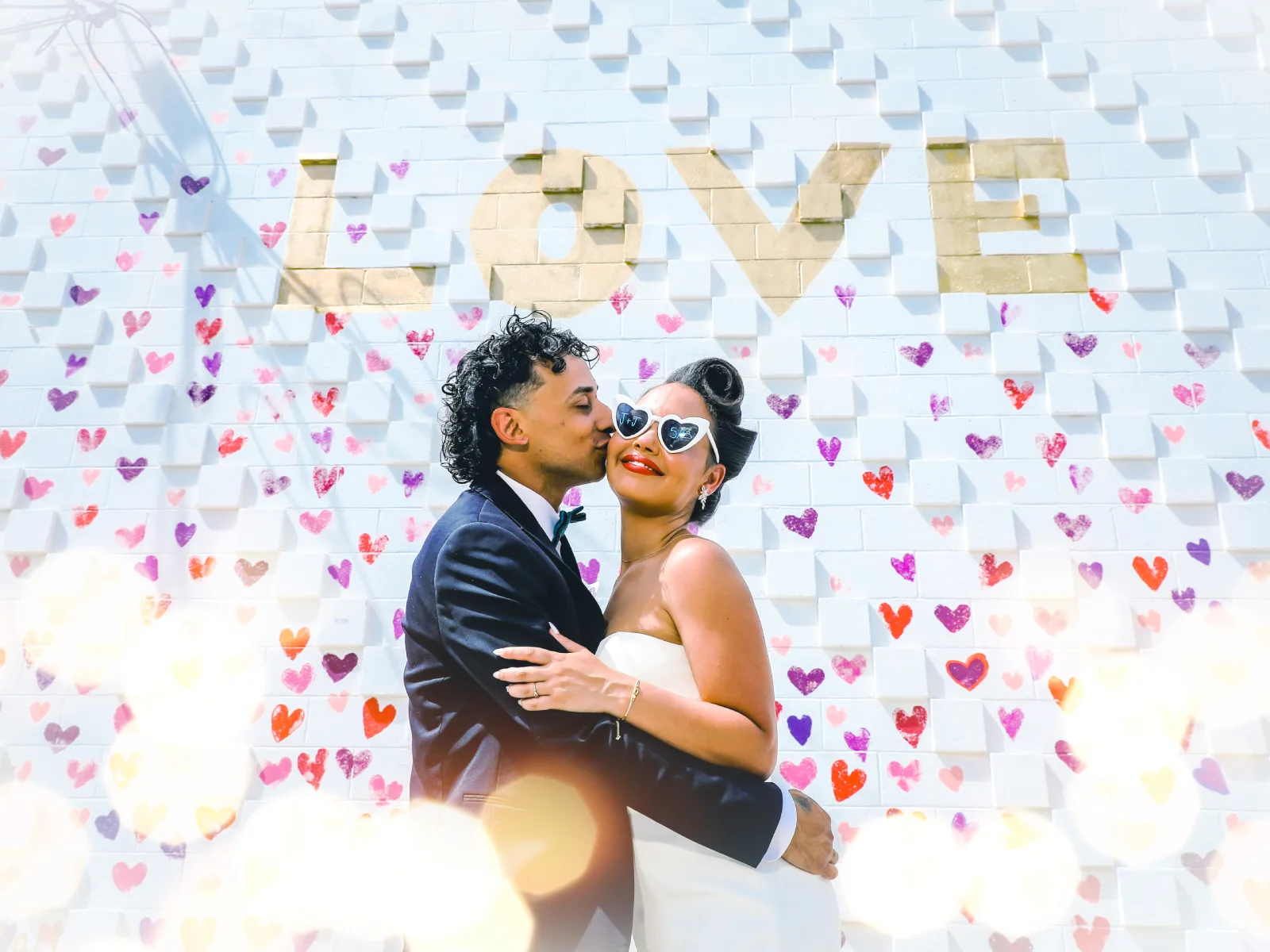A playful groom kissing the cheek of a bride wearing heart-shaped sunglasses, standing against a white brick wall decorated with colorful hearts and a "LOVE" sign.