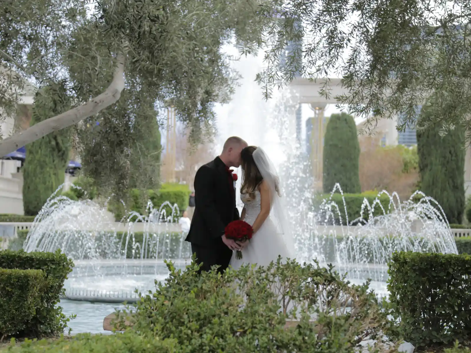A bride and groom sharing a romantic kiss in front of a grand water fountain, framed by lush greenery and olive trees.
