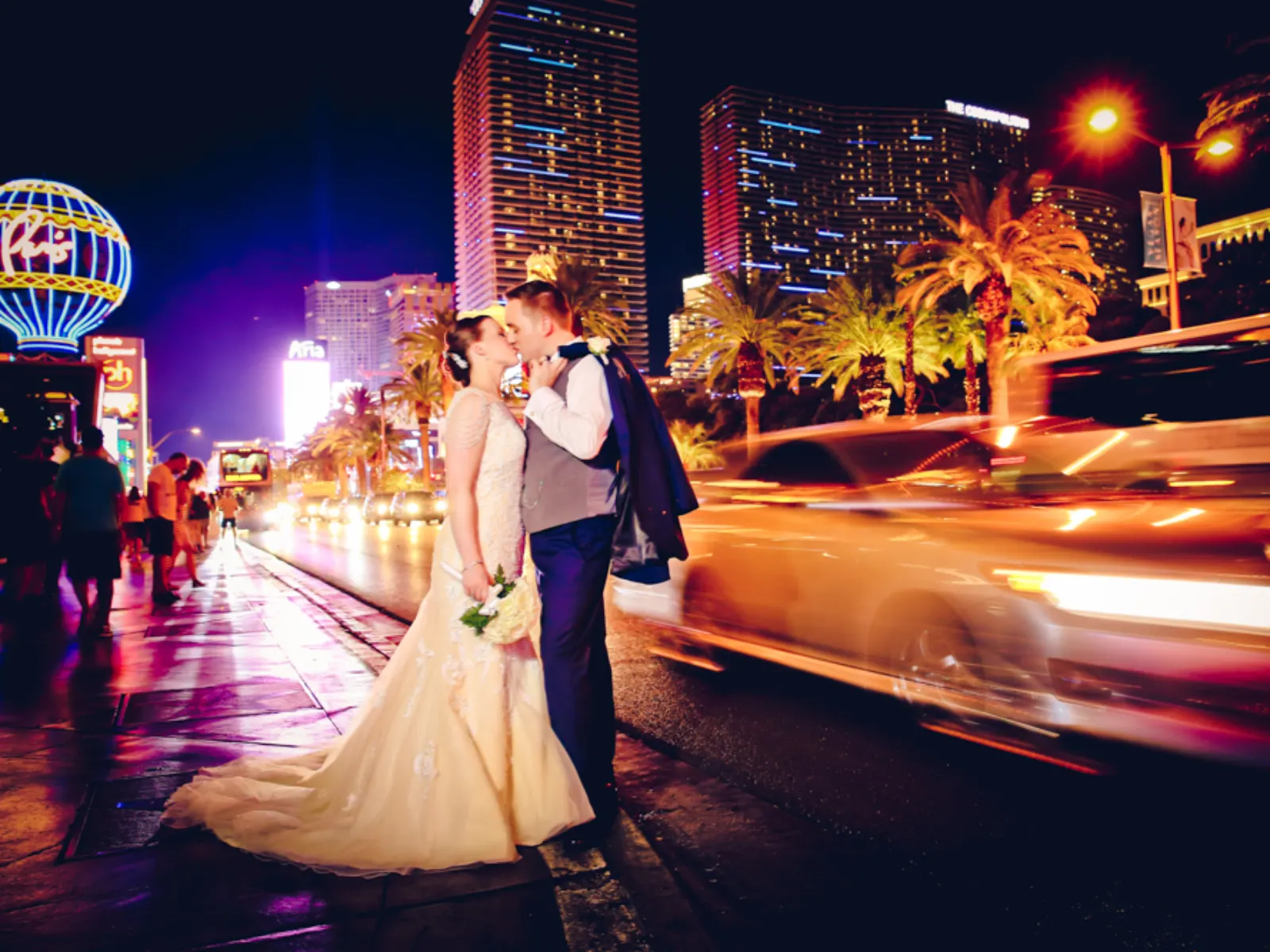 A bride and groom kissing on the Las Vegas Strip at night, with bright city lights, palm trees, and the motion blur of a passing car in the background.