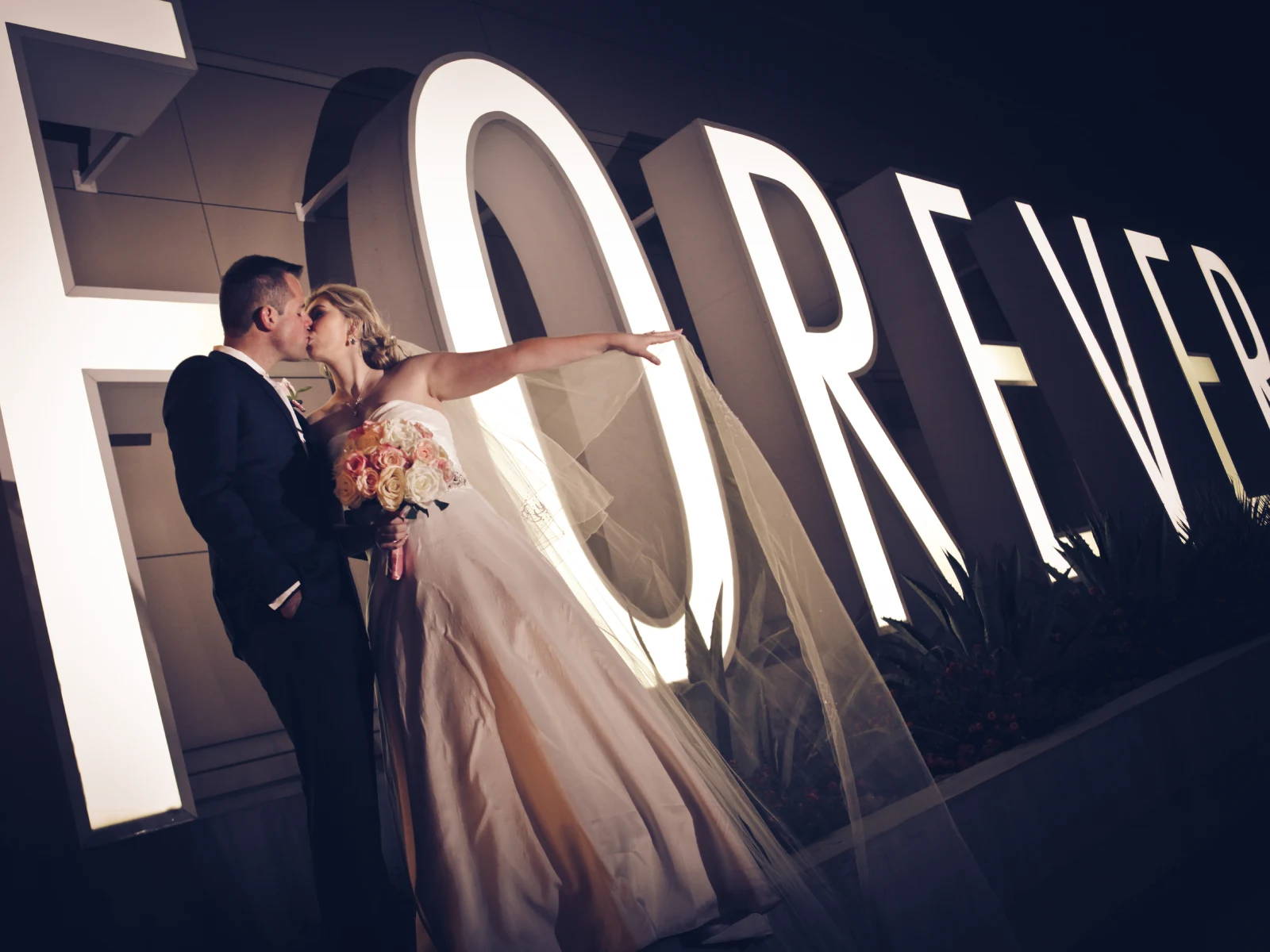 A bride and groom kissing at night in front of a large, glowing white "FOREVER" neon sign with a dramatic low-angle perspective.