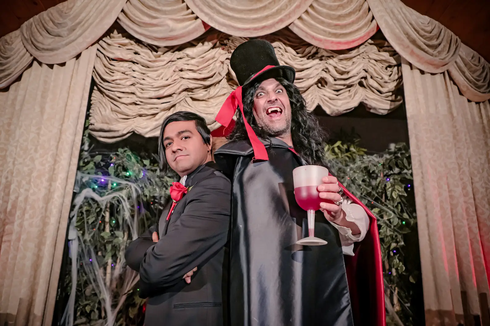 Groom posing back to back with Dracula impersonator in cape and top hat holding red goblet at Las Vegas chapel