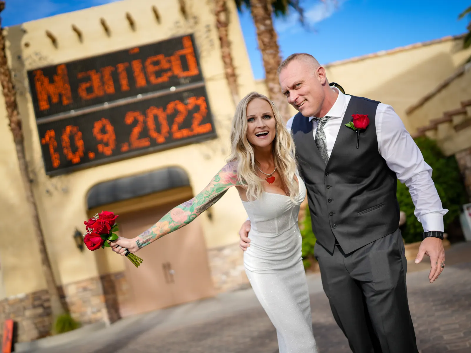 A happy bride with a floral sleeve tattoo and a groom in a charcoal vest walking in front of a Las Vegas wedding chapel with a marquee sign reading "Married 10.9.2022."