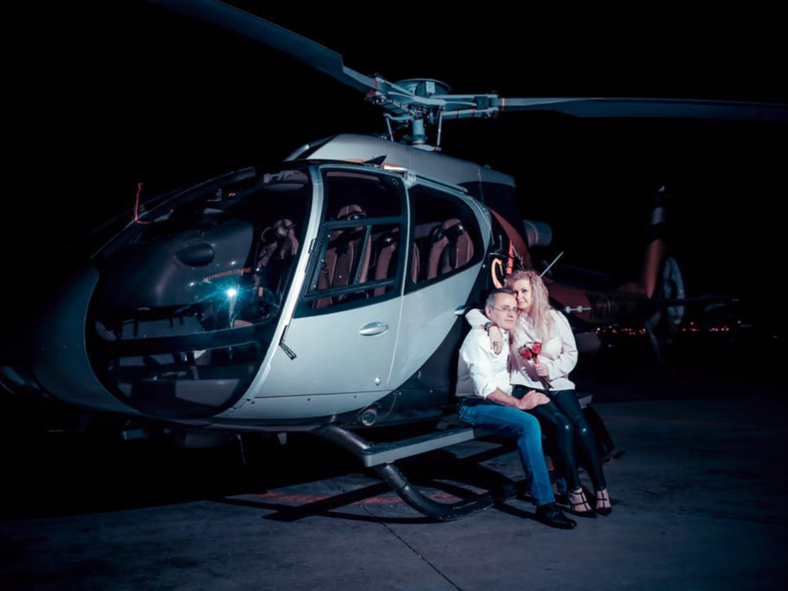A couple in semi-formal attire sitting on the landing skid of a silver helicopter at night, with the distant glow of the Las Vegas city lights in the background.