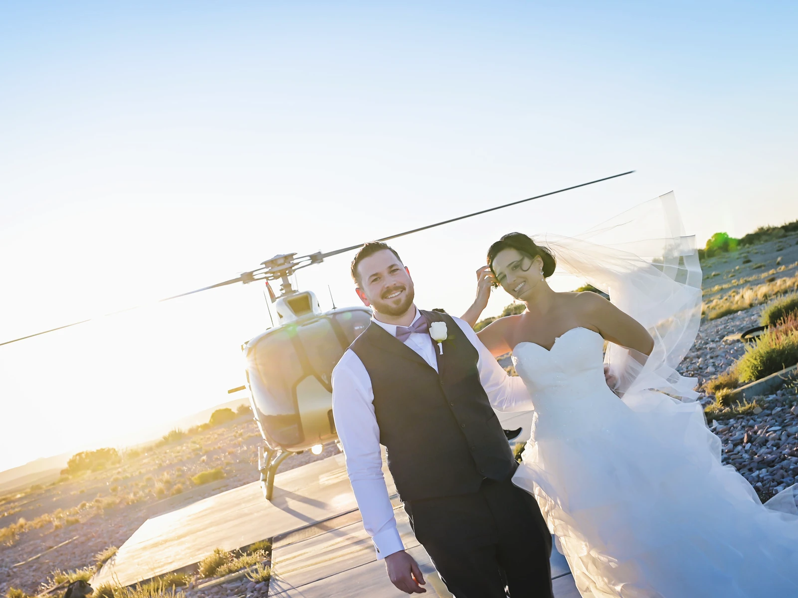 A close-up, focused shot of a vibrant pink rose and lily bouquet resting on a gravel ground, with a blurred bride and groom embracing in front of a white wedding gazebo in the background.