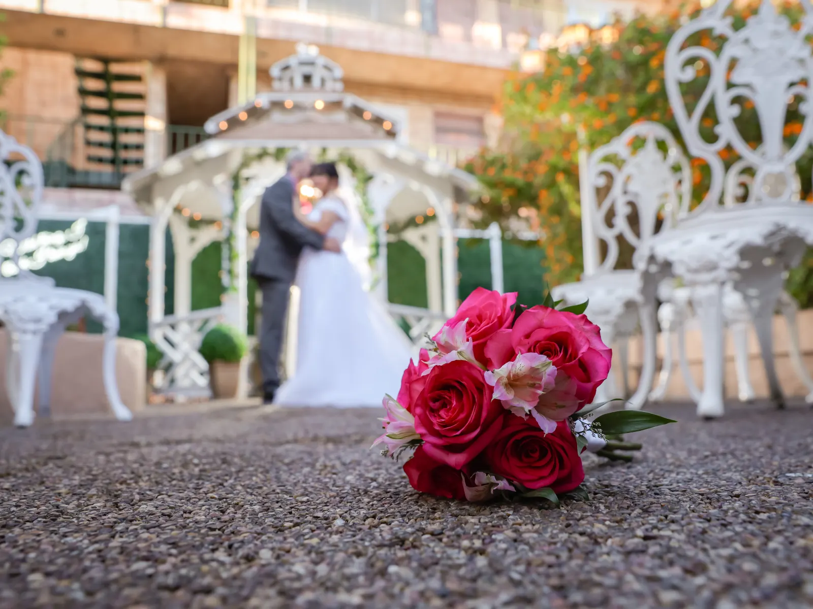 A close-up, focused shot of a vibrant pink rose and lily bouquet resting on a gravel ground, with a blurred bride and groom embracing in front of a white wedding gazebo in the background.